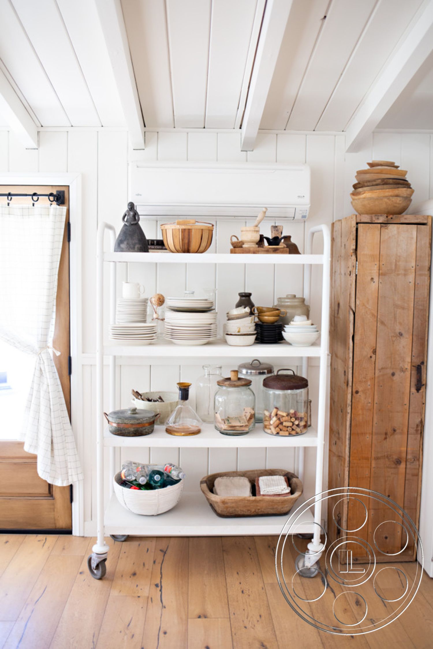 Ranch 33 - neutral painted walls, textured linen curtains, minimalist kitchen aesthetic, beadboard wall treatment, light oak hardwood flooring, interior shelving unit with white metal frame, rustic wood cabinet with natural finish, high-gloss ceramic dishware, clear glass storage jars with metal lids, bamboo serving bowls, natural wood cutting board, simple white porcelain plates, matching black ceramic cups, artisanal pottery, small round wicker basket, decorative wooden sculptures, open shelving design for kitchen display, contemporary farmhouse style, functional kitchen storage solution, organized clutter display