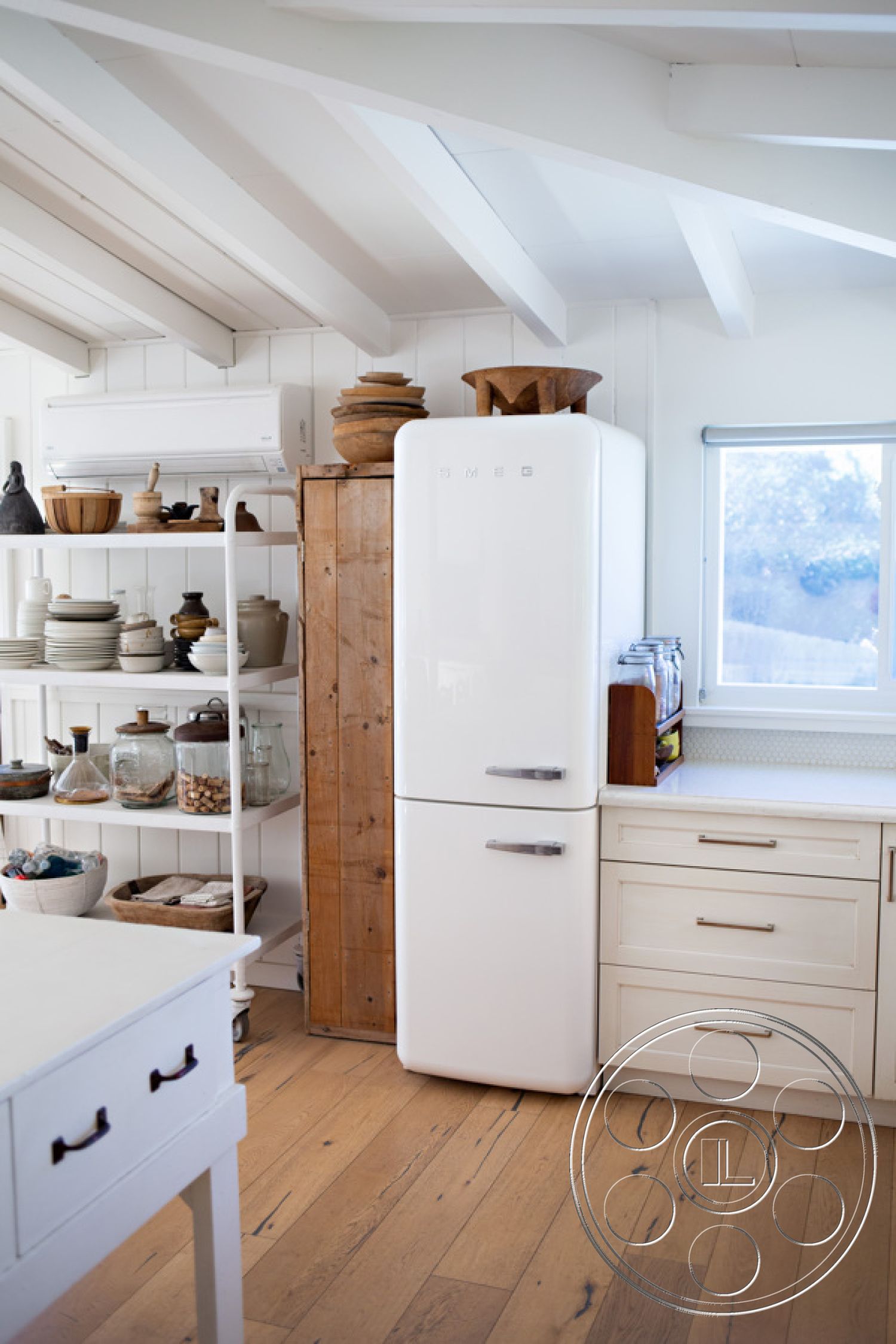 Ranch 33 - functional kitchen space, modern farmhouse style, natural light from window, glass storage jars, exposed wood ceiling beams, minimalist kitchen design, hardwood kitchen flooring, white painted walls, open shelving unit, dining area with wooden table, Smeg refrigerator, rustic wooden cabinet, light-colored countertops, artistically arranged kitchen utensils
