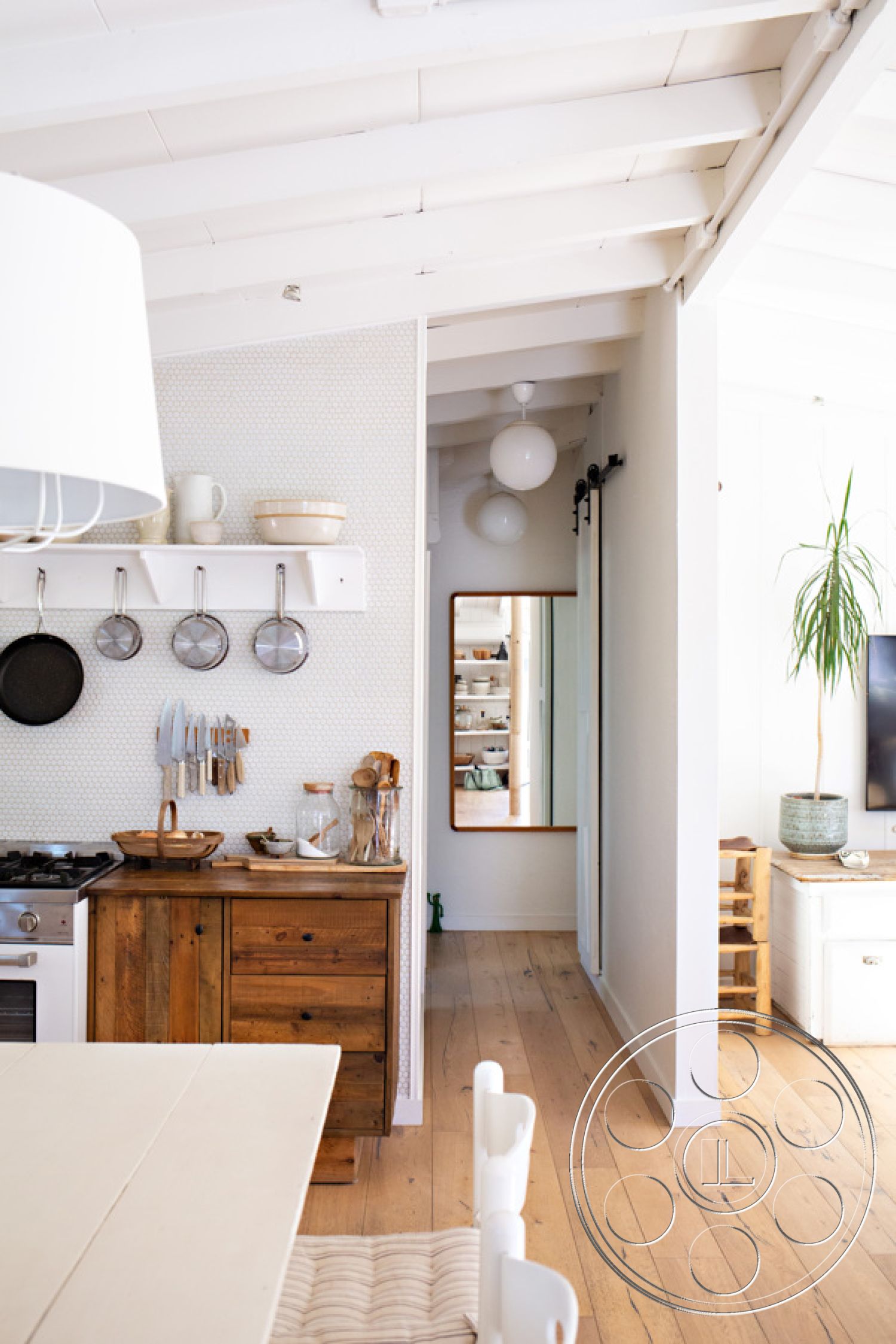 Ranch 33 - neutral color palette, modern farmhouse style, exposed wood ceiling beams, stainless steel kitchen appliances, wooden kitchen cabinets, open-concept dining area, hardwood kitchen floors, textured wallpaper accent wall, white painted interior walls, minimalist home decor, natural light from surrounding windows, Pendant light fixture above dining table, large wall mirror in hallway, shelves with dishware in kitchen, plant decor in living area