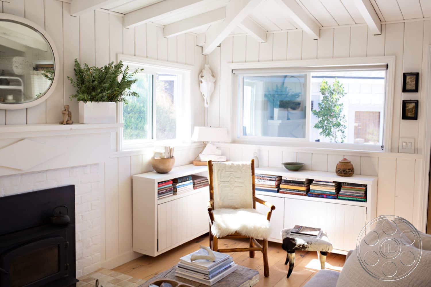 Ranch 33 - interior neutral color palette, interior vaulted ceiling with exposed wood beams, exterior white shiplap walls, interior bright natural light from multi-pane windows, interior white brick fireplace, interior hardwood floor in warm tones, interior built-in white cabinetry, interior round mirror as wall decor, interior cozy reading nook with plush chair, interior decorative potted plant on windowsill, interior soft area rug on hardwood flooring, exterior garden view visible through windows