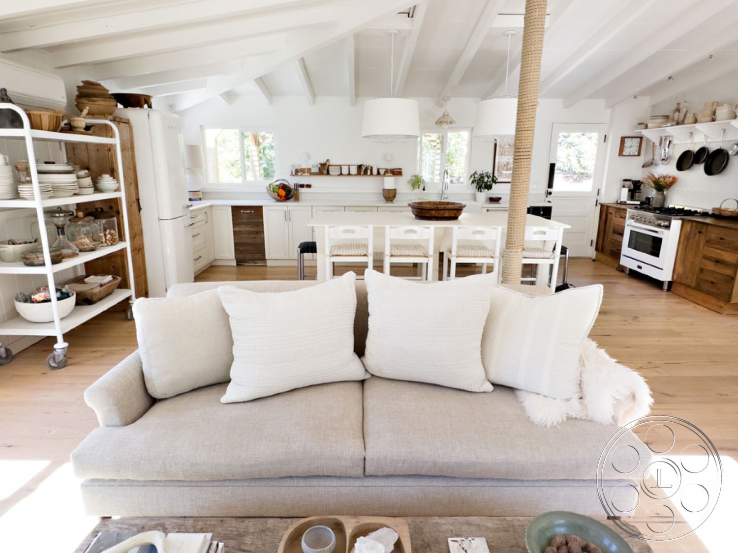 Ranch 33 - open concept layout, exposed wood ceiling beams, textured throw pillows on sofa, white painted walls in kitchen, light hardwood flooring in living room, modern kitchen with wood cabinetry, glass pendant lighting fixtures, natural stone kitchen countertops, dining area with natural wood table, cozy seating arrangement with soft fabrics, sunlit interior with ample windows, minimalist decor with functional shelving, interior garden view from windows