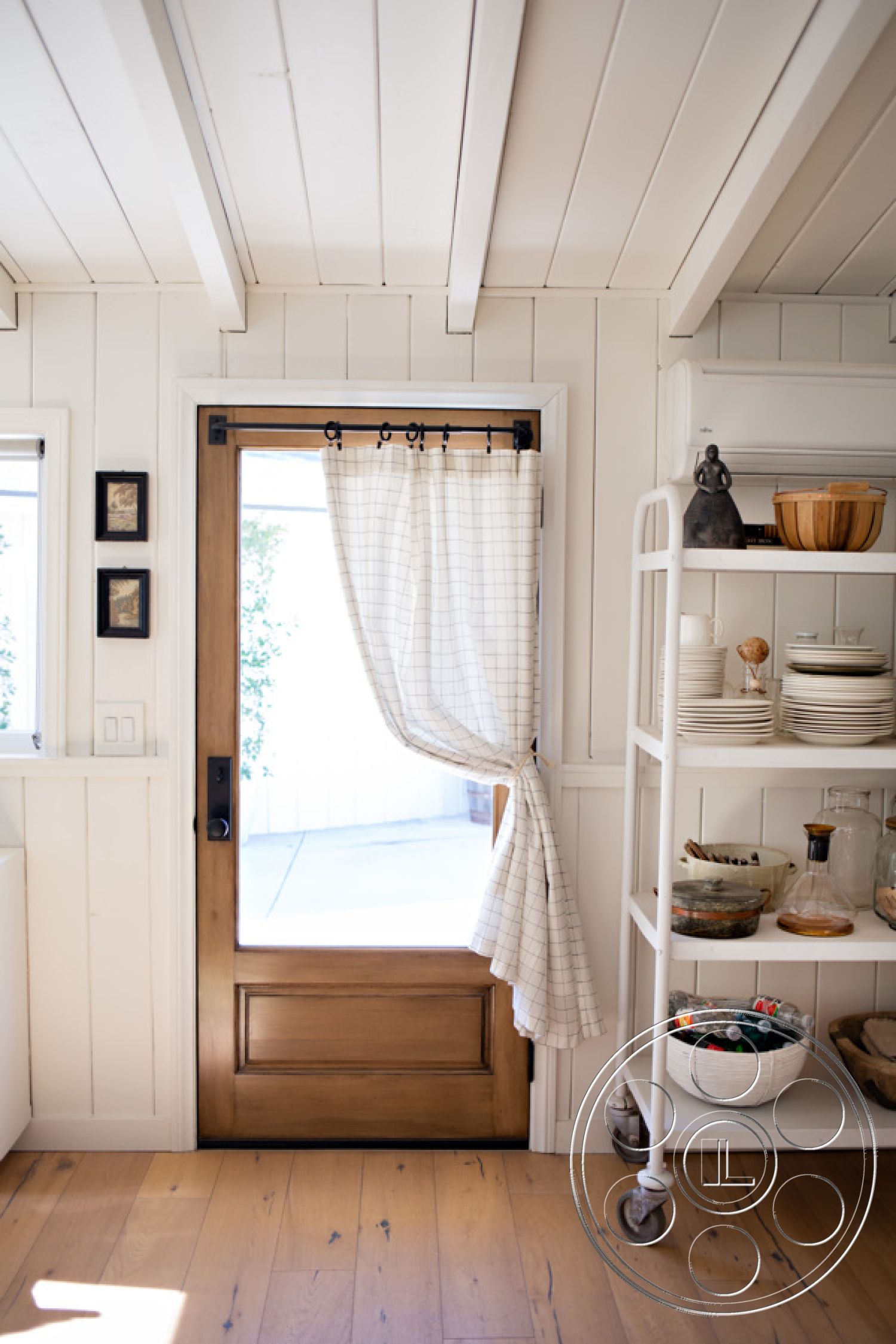 Ranch 33 - casual dining atmosphere, rustic decor elements, functional kitchen layout, minimalist kitchen design, open shelving kitchen storage, light-filled interior space, natural hardwood flooring, white shiplap wall treatment, interior wood door with glass insert, black accent frames on white walls, textured fabric curtain with grid pattern, exposed wood beams on ceiling, modern farmhouse entryway, bright room with natural light, contemporary kitchen accessories