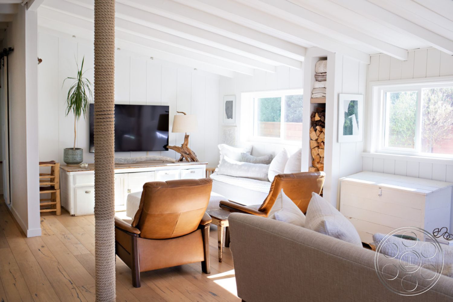 Ranch 33 - neutral color palette, hardwood flooring, cozy seating area, white shiplap walls, textured throw pillows, natural light from large windows, exposed wood ceiling beams, interior living room, minimalist decor accents, decorative potted plant, rustic wooden coffee table, modern flat-screen television, comfortable leather armchair, white couch with cushions, built-in bookshelf with firewood