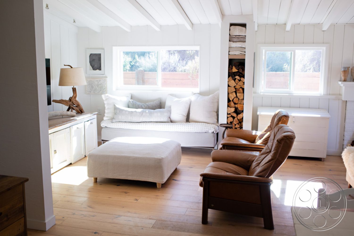 Ranch 33 - hardwood floor living room, neutral color palette interior, exposed wood ceiling beams interior, natural light-filled space interior, modern lighting fixtures interior, large double-pane windows, minimalist decor interior, open concept interior space, cozy living room design, white shiplap walls interior, plaid upholstered sofa, leather armchairs interior, square upholstered ottoman, built-in wood storage for logs