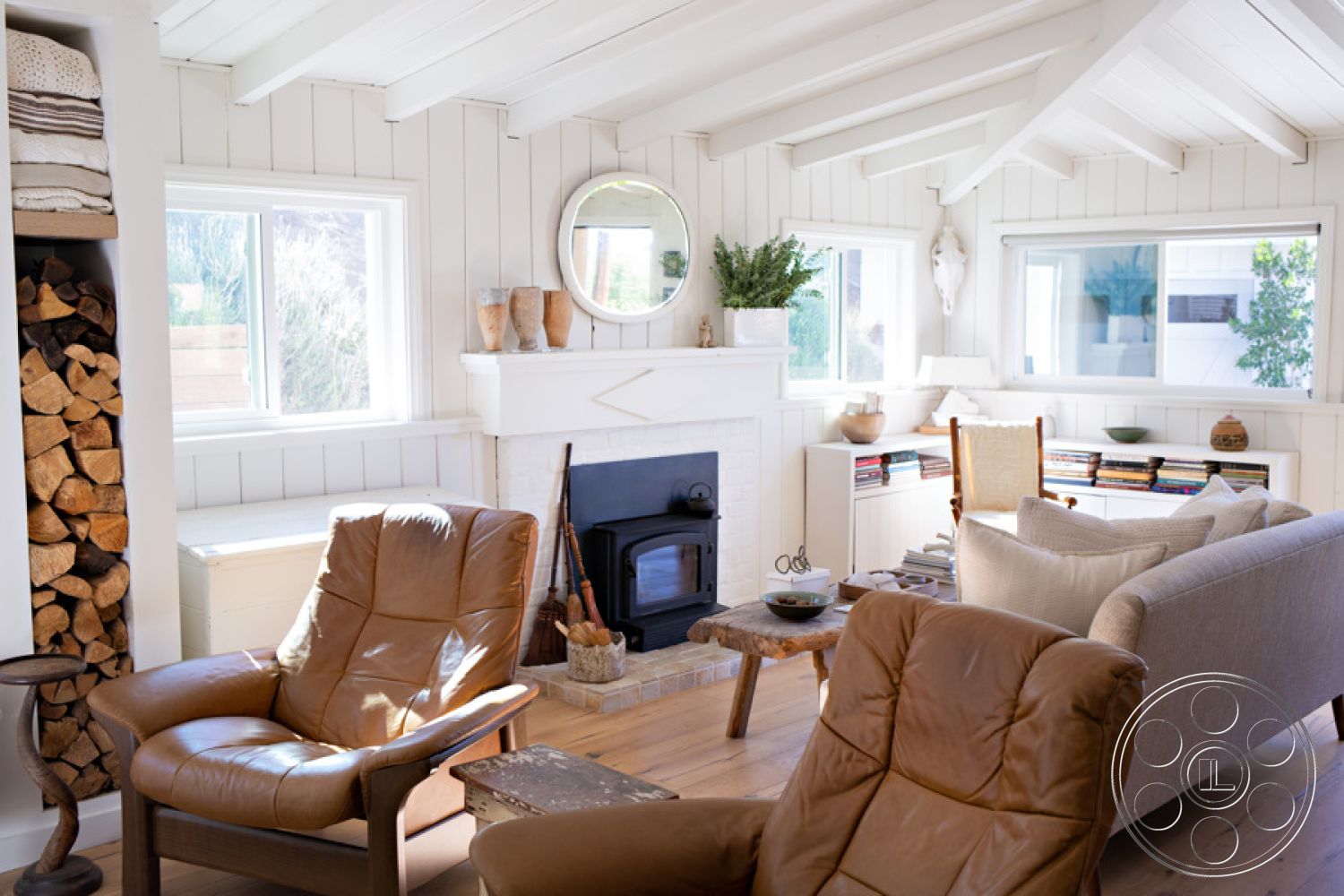 Ranch 33 - exposed wood ceiling beams interior, large windows with natural light interior, hardwood flooring living room interior, stone fireplace surround interior, white shiplap walls interior, dark leather armchairs interior, natural wood coffee table interior, built-in bookcases interior, simple round mirror wall decor interior, decorative plant accents interior, stacked wood storage interior, simple shelving unit interior