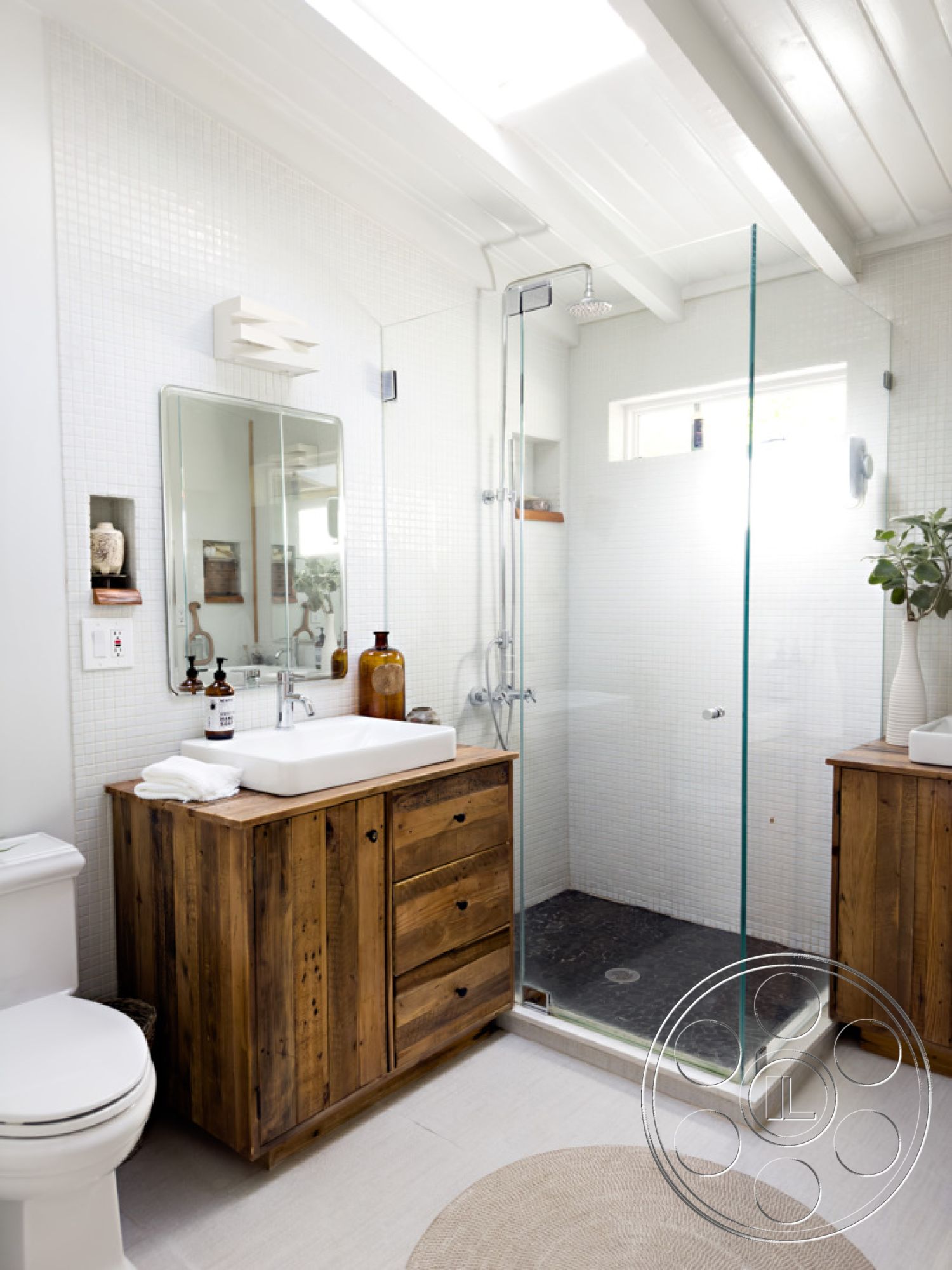Ranch 33 - neutral color palette, minimalist design, glass shower enclosure, open space layout, potted greenery, white porcelain toilet, chrome shower fixtures, interior bathroom, white shiplap ceiling, modern rectangular sink, reclaimed wood vanity with drawers, black stone shower floor, white mosaic tile shower walls, natural fiber round rug, window for natural light, decorative glass bottle accents