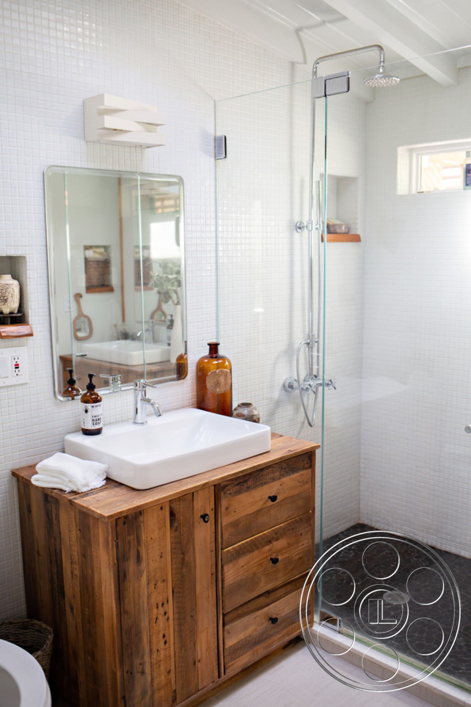 Ranch 33 - neutral color palette, glass shower enclosure, eco-friendly materials, natural light from window, interior bathroom design, chrome shower fixtures, minimalist bathroom decor, modern wall-mounted light fixture, white mosaic tile walls, handcrafted wooden vanity, rustic wood bathroom cabinet, square porcelain sink, black pebble stone shower floor, floating shelves for bathroom storage, mirror with wooden frame, soft white linen towel, open-concept shower area, refined rustic aesthetic