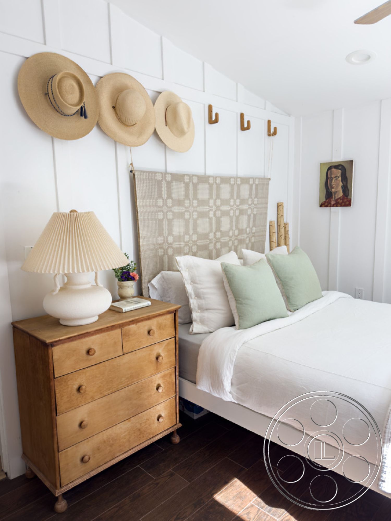 Ranch 33 - green accent pillows, woven wall hanging, decorative flower pot, interior bedroom, hardwood bedroom floors, textured fabric headboard, white painted walls, beige upholstered bedding, wooden dresser with six drawers, table lamp with round base, straw hats mounted on wall hooks, natural light from ceiling fixture