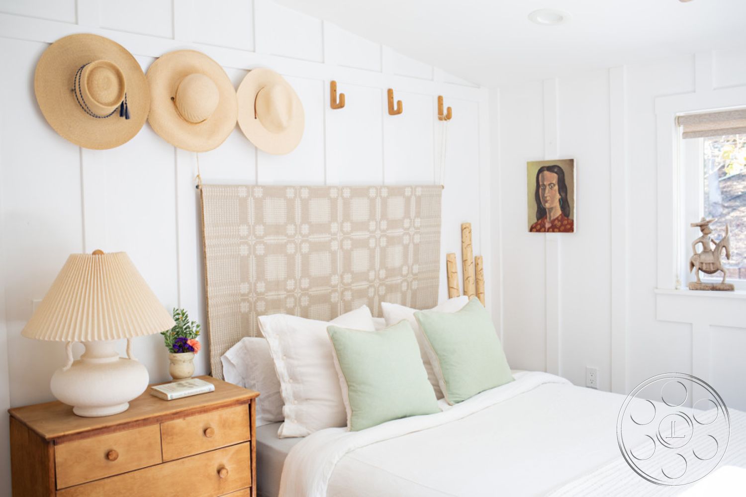 Ranch 33 - wooden bedside table, quilted bedspread, green accent pillows, natural light from window, interior bedroom, small potted plant decor, white shiplap wall treatment, textured woven wall hanging, natural wood hooks, ceramic lamp with fabric shade, framed portrait artwork, subtle rustic decor