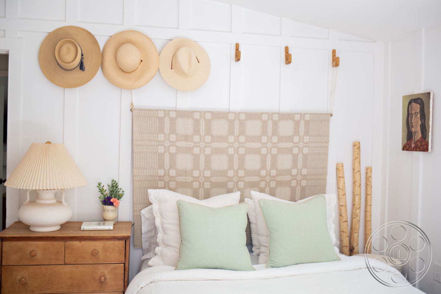 Ranch 33 - interior bedroom with shiplap wall treatment, fabric-covered headboard with geometric pattern, hardwood floor in bedroom, pastel green throw pillows on bed, decorative lamp with pleated shade, wooden nightstand with drawers, dried flower arrangement in ceramic vase, straw hats displayed on wall hooks, natural wood accents in bedroom decor, artwork on wall with portrait, birch logs as decor element