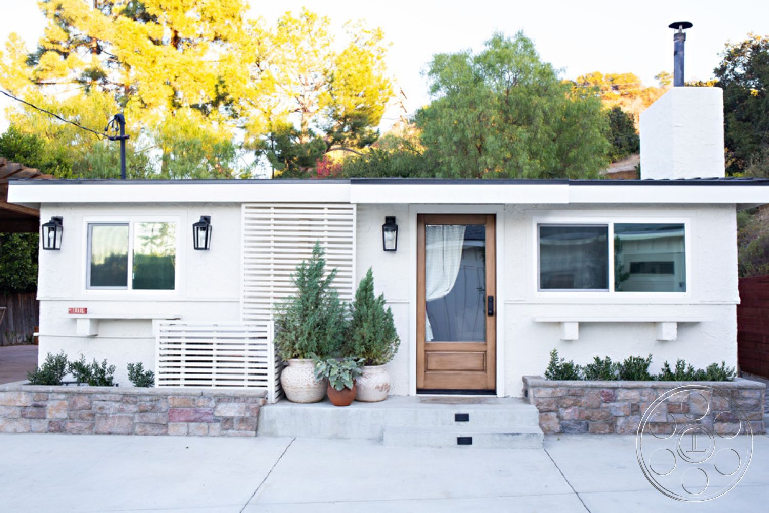 Ranch 33 - exterior, white stucco walls, single-story home, surrounded by trees, wood front door, hillside location, stone planters, flat roof design, modern minimalist aesthetic, wooden porch overhang, exposed stone entry wall, concrete landing steps, rectangular picture windows, black lantern wall sconces, horizontal wooden slats screen, desert garden landscaping, potted succulents and shrubs