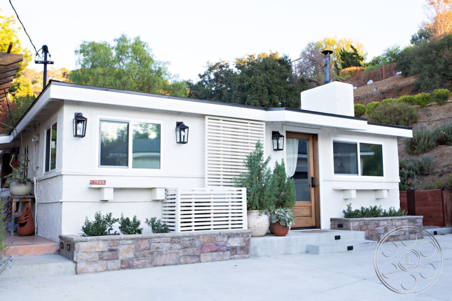 Ranch 33 - exterior, stucco finish walls, exterior, flat roof design, exterior, stone planter border, exterior, concrete driveway, exterior, wooden front door, exterior, horizontal wooden slats, exterior, black wall-mounted lanterns, exterior, landscaped garden beds, exterior, low-maintenance shrubs, exterior, hillside location, exterior, modern architectural style