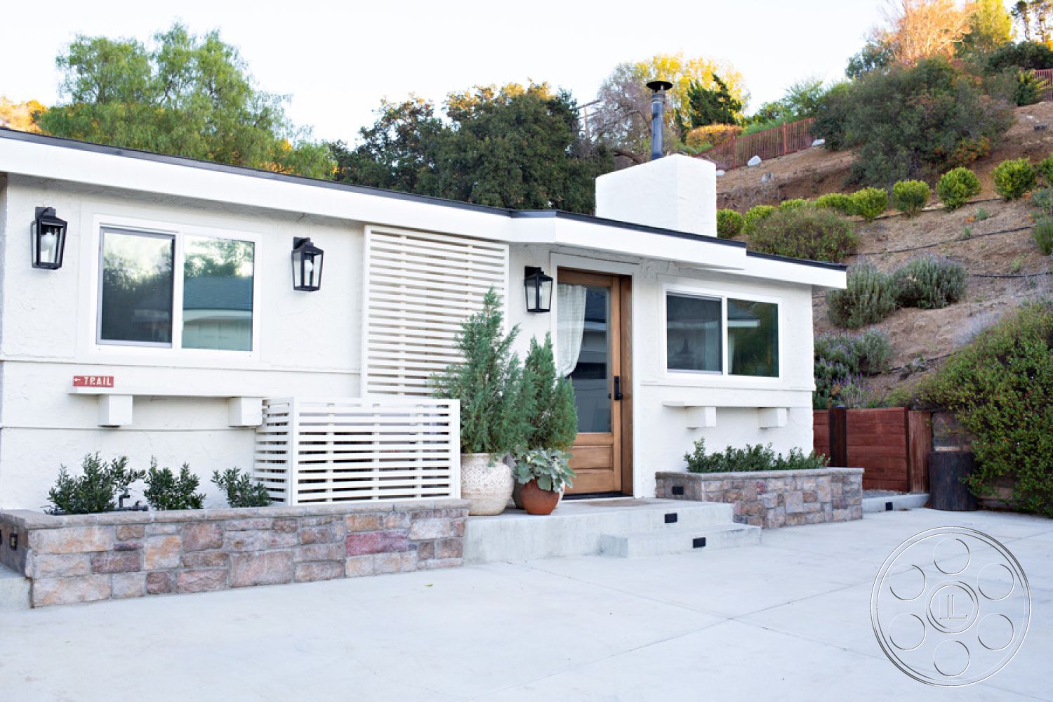 Ranch 33 - exterior, natural stone accents, landscaped hillside, stucco walls, wooden front door, flat roof design, concrete patio flooring, stone flower bed border, urban hillside setting, wall-mounted outdoor lighting, double-pane windows, concrete step entrance, decorative horizontal slats, shrubs in garden beds