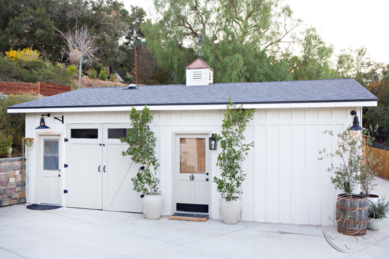 Ranch 33 - exterior, asphalt shingle roof, natural wood accents, landscaped garden area, wooden double doors, white vertical siding, stone foundation walls, decorative garage door with windows, black metal wall sconces, cement driveway, potted greenery at entrance, continuous ridge vent, light fixture with glass shade, sloped lot setting, rustic barn-style elements, wooden planter barrels, cement pathway leading to door