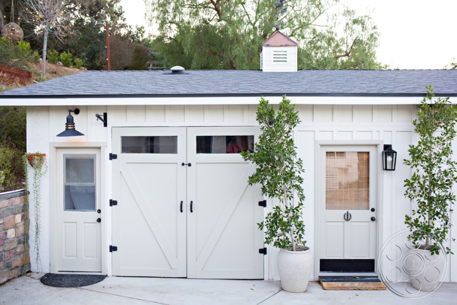 Ranch 33 - neutral color palette, exterior, slate roof, minimalistic aesthetic, functional outdoor storage, stone garden wall, double door entrance, wooden barn-style doors, white board and batten siding, landscaped with potted plants, modern light sconces, small side door, organized outdoor space