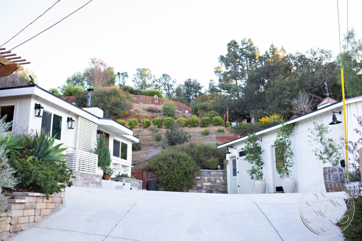 Ranch 33 - concrete driveway, white painted exterior walls, exterior lighting fixtures, landscaped hillside garden, sloped terrain setting, modern farmhouse architectural style, exposed stone retaining wall, horizontal wood slat exterior details, garden planters with greenery, vegetation accents in yard, urban-rural transitional setting, patio access from main entrance, outdoor landscaping elements, natural aesthetic with surrounding trees, minimalist geometric shapes