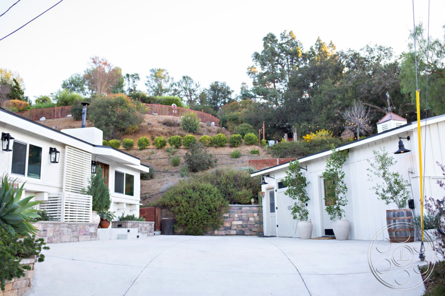Ranch 33 - concrete patio, landscaped hillside, hilltop view, concrete driveway, exposed light fixtures, stone retaining wall, textured landscape, landscaped entryway, modern ranch-style home, outdoor garden area, planter pots with greenery, exterior white wood siding, desert home setting, surrounding natural vegetation, driveway lead-in