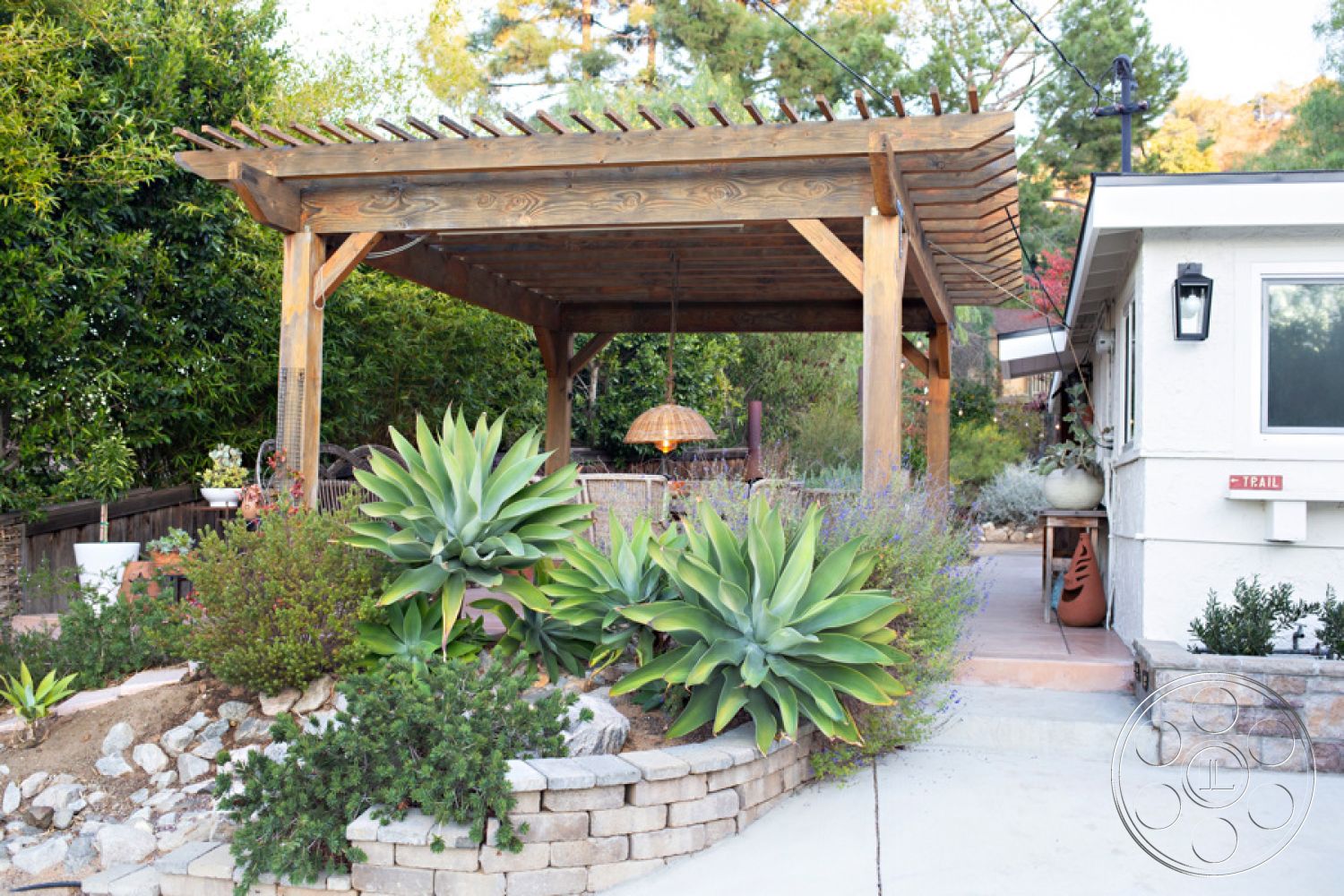 Ranch 33 - natural stone retaining wall, modern outdoor living space, cozy outdoor seating area, lush green foliage backdrop, wood pergola structure exterior, ceramic tile patio flooring exterior, stucco house wall exterior, landscaped garden area exterior, paved stone garden border, hanging decorative light fixture, metal outdoor lantern light fixture, succulent and agave plants landscaping, concrete boardwalk pathway exterior, wooden outdoor dining furniture, rustic wooden beam design, open-air relaxation space exterior, tranquil garden setting exterior, crafted outdoor plant arrangements