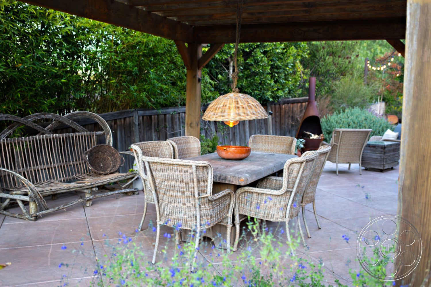 Ranch 33 - slate stone flooring, outdoor fireplace, decorative light fixture, exposed wood ceiling beams, wood beam pergola, landscaped garden setting, exterior patio area, rustic outdoor seating, seasonal flowering plants, wooden fence enclosure, woven rattan dining chairs, weathered wood dining table, ceramic bowl centerpiece, bamboo plant accents, suburban backyard dining space