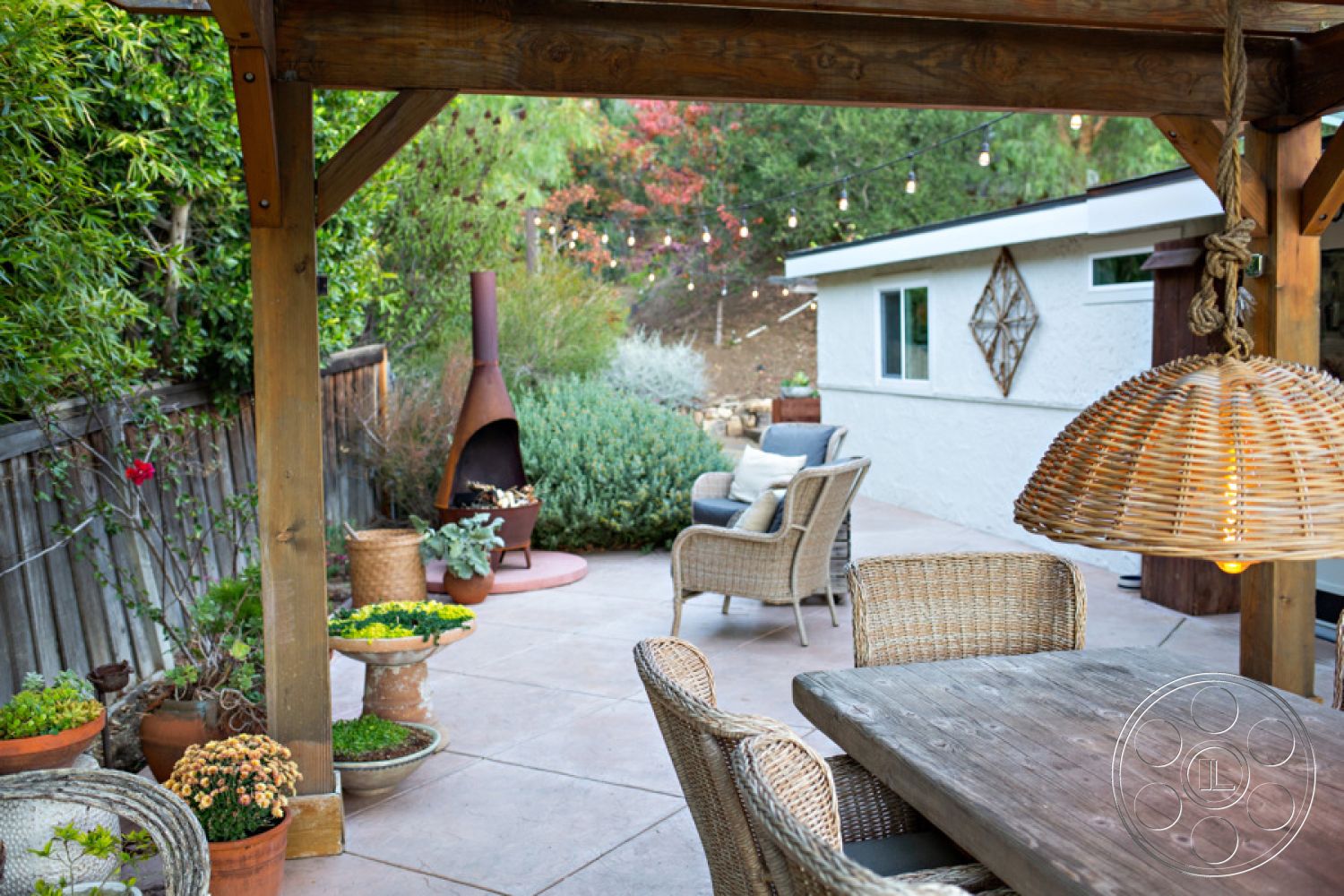 Ranch 33 - natural wood accents, ambient string lights, lush greenery backdrop, landscaped garden setting, wood pergola structure, exterior patio area, rustic wooden dining table, woven rattan dining chairs, open-air dining experience, outdoor fireplace feature, stamped concrete patio flooring, ceramic potted succulents, hanging woven light fixture, modern home with white stucco walls, round decorative plant stands, backyard relaxation space