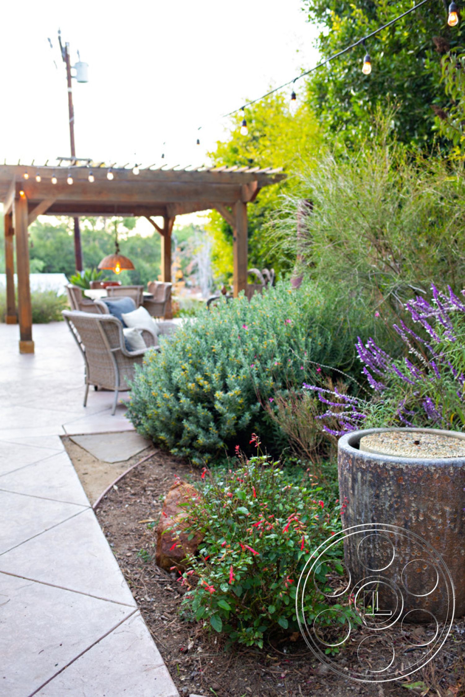 Ranch 33 - natural stone elements, colorful flowering plants, sustainable landscaping, relaxed outdoor atmosphere, modern outdoor seating, landscaped garden beds, exterior patio area, rustic garden setting, lush greenery surrounding, wooden pergola structure, string lights overhead, wicker patio furniture, textured concrete patio floor, decorative stone water feature, earthy stone accent boulders, alleyway access landscape design