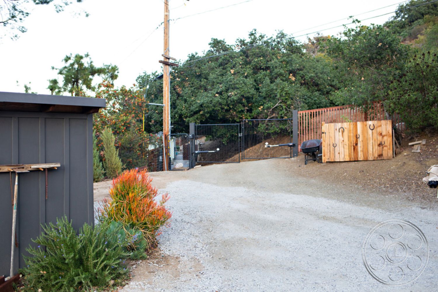 Ranch 33 - outdoor seating area, exposed wooden beams, mountain backdrop, rural setting, gravel driveway, landscaped garden with shrubs, decorative wooden accents, metal mesh fencing, textured wood siding, open hillside view, outdoor grill area, pergola structure, wooden fence gate, heavily wooded area, rustic wooden storage shed, natural stone landscape, gray gravel surface, steep incline driveway, neighbors' trees in background, electric utility pole