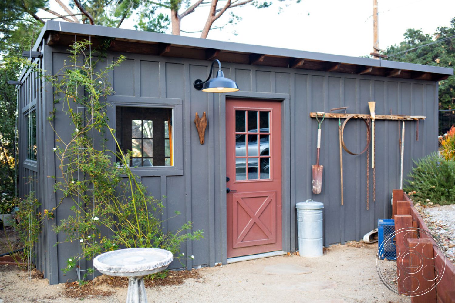 Ranch 33 - rustic outdoor decor, scenic garden setting, natural light source, exterior wooden shed, gray vertical wood siding, decorative red panel door, single-glazed windows, metal gooseneck light fixture, exposed wooden roof beams, landscape climbing plants, metal garden tools on wall, gravel yard surface, circular birdbath feature, surrounding brush landscape, farmhouse style shed, simple rustic architecture, functional garden storage, low-maintenance exterior