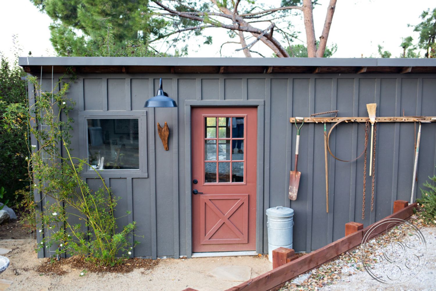 Ranch 33 - decorative garden elements, exterior wood paneling, landscaped gravel area, rustic outdoor aesthetic, casual backyard setting, dark gray wood siding, red wooden front door with glass panes, metal barn light fixture, small square window, concrete path leading up to the entrance, wooden garden tools rack, surrounding greenery and plants, open sky above, modern garden shed design
