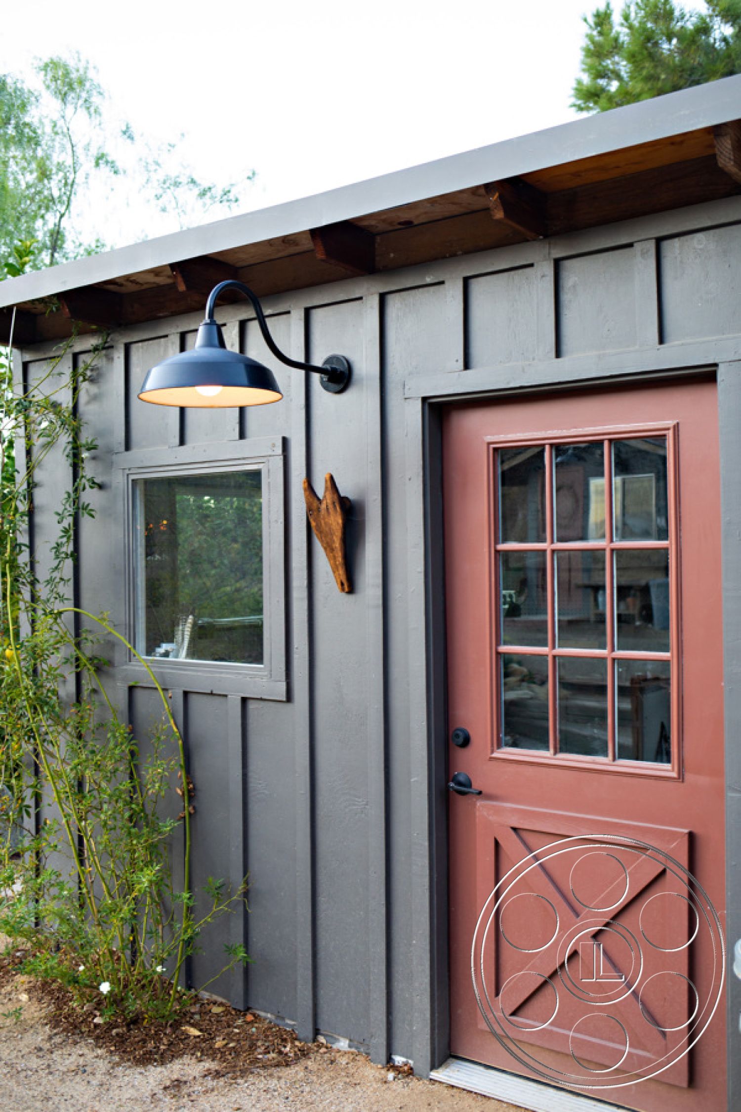 Ranch 33 - minimalist design, natural wood trim, wood siding exterior, gray wood paneling, red painted entrance door, double-paned window with wood frame, metal wall-mounted light fixture, exposed rafters on roof, landscape with shrubs and plants, vertical wood siding textures, rustic exterior accents, outdoor lighting details, country-style architecture, garden path leading to entrance