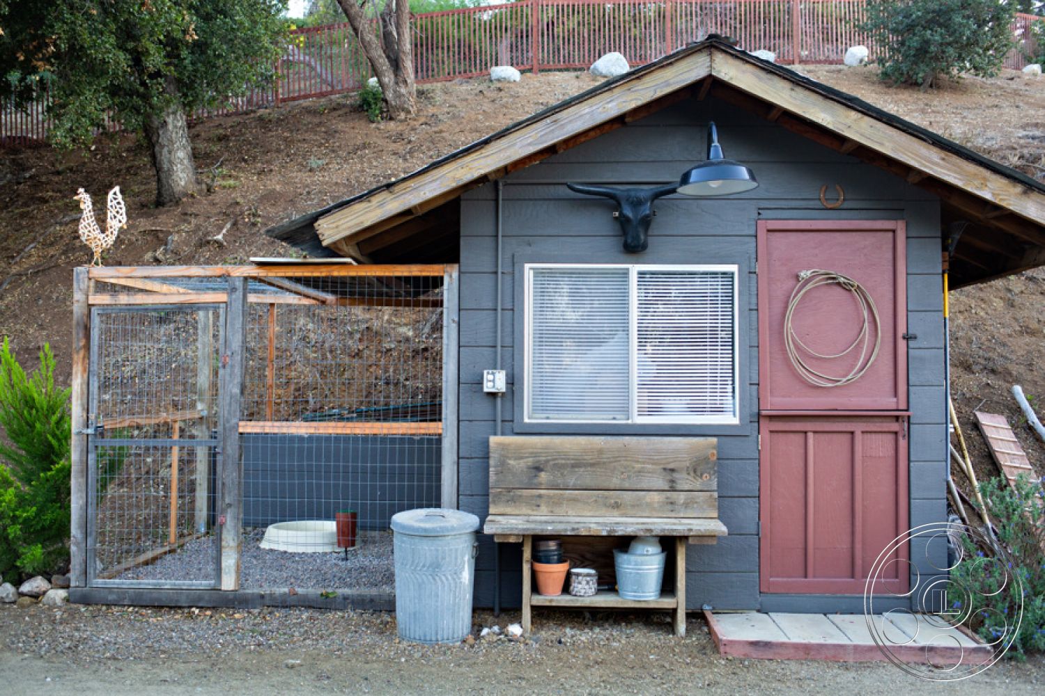 Ranch 33 - metal light fixture, wooden garden bench, stone patio flooring, landscaped hillside setting, wood beam porch ceiling, rural farmhouse style, gabled roof structure, gravel driveway surface, wooden exterior siding, exposed wooden porch railing, red painted front door, metal animal enclosure, interior wooden window framing, rustic window blinds