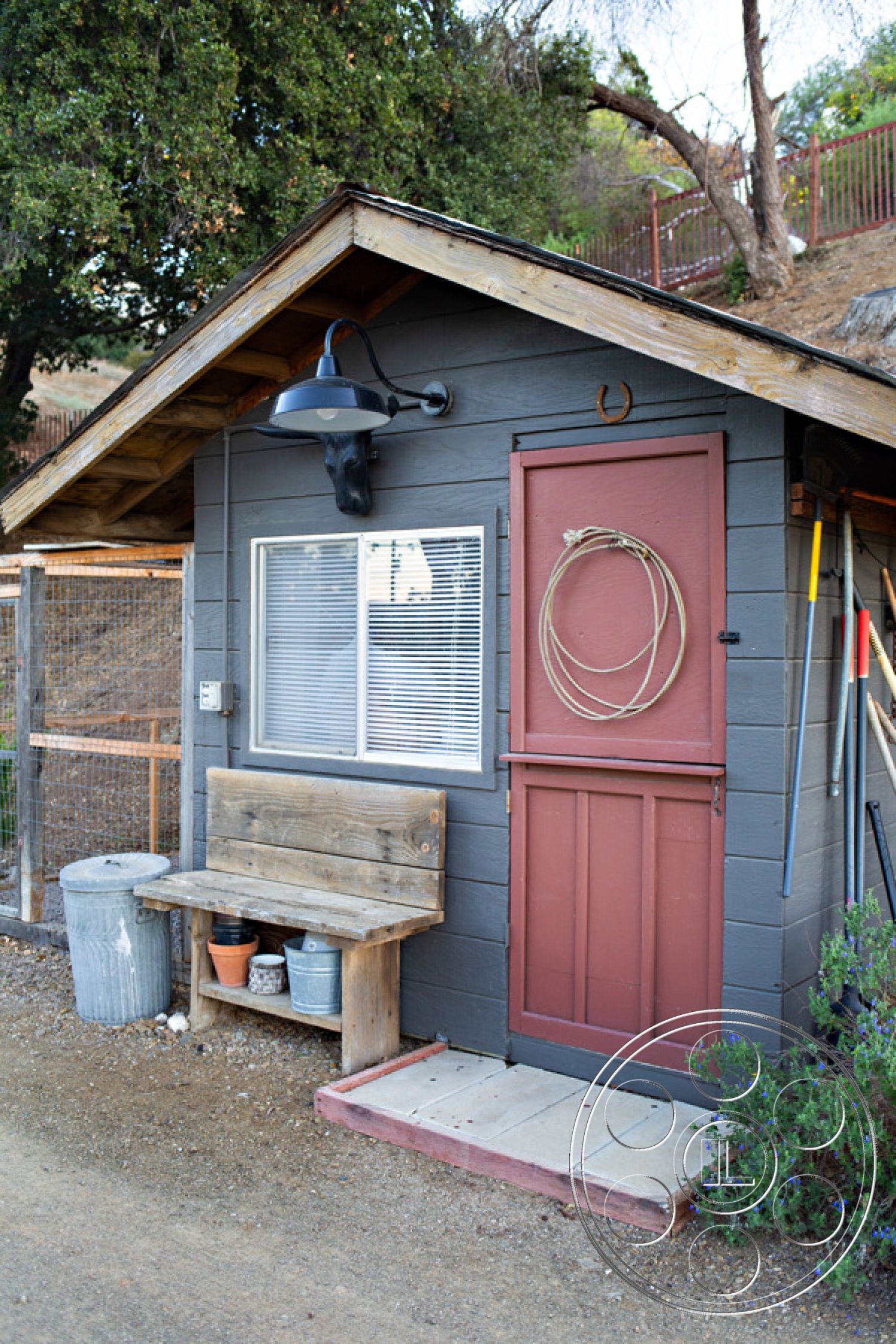 Ranch 33 - farmhouse style, mountainous backdrop, metal light fixture, natural stone walkway, landscaped garden with shrubs, glass window with white trim, exterior wooden cabin, exposed wooden framework, gravel patio area, shiplap gray siding, wooden bench with rustic finish, red wooden door, rustic rural setting, wooden ranch-style roof overhang, decorative rope wall hanging, functional outdoor storage space