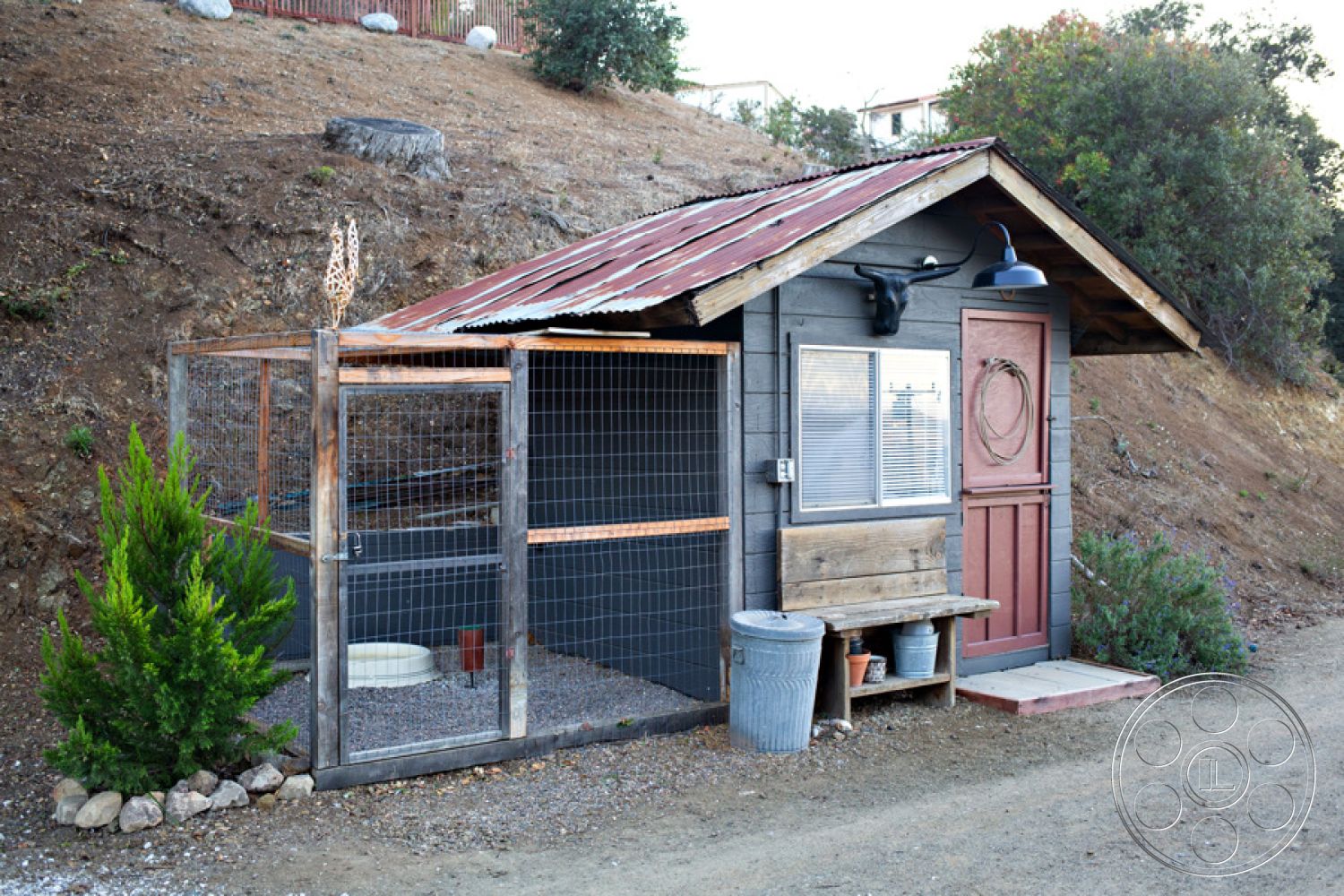Ranch 33 - glass window features, mountain hillside setting, country-style architecture, wooden exterior walls, metal roof with rustic finish, wooden bench seat, wire mesh enclosure, gravel flooring in animal area, exposed wooden frame structure, red barn-style door, outdoor pet enclosure, weathered wood accents, decorative wall light fixture, landscaped with native vegetation, interior animal comfort area, outdoor utility space