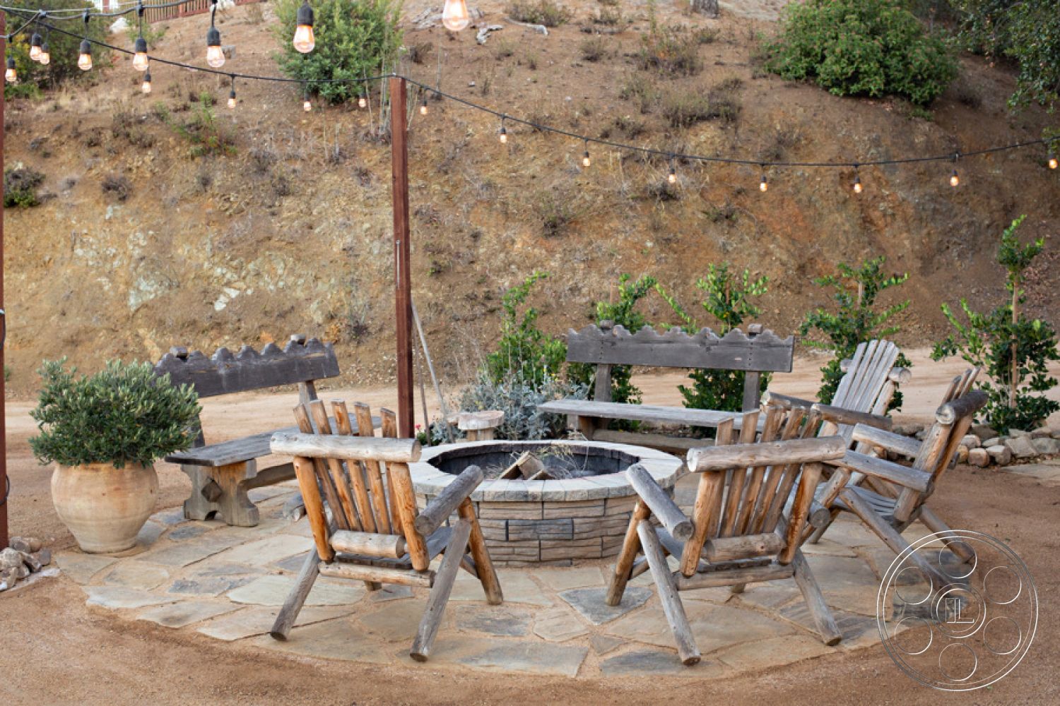 Ranch 33 - gravel pathway, wooden bench seating, natural stone pavers, ambient string lighting, exterior stone patio flooring, natural hillside setting, round stone firepit, weathered wood Adirondack chairs, landscape planter pots, desert home outdoor retreat, rustic outdoor gathering area, wood post structures, drought-resistant shrubs, relaxed backyard environment