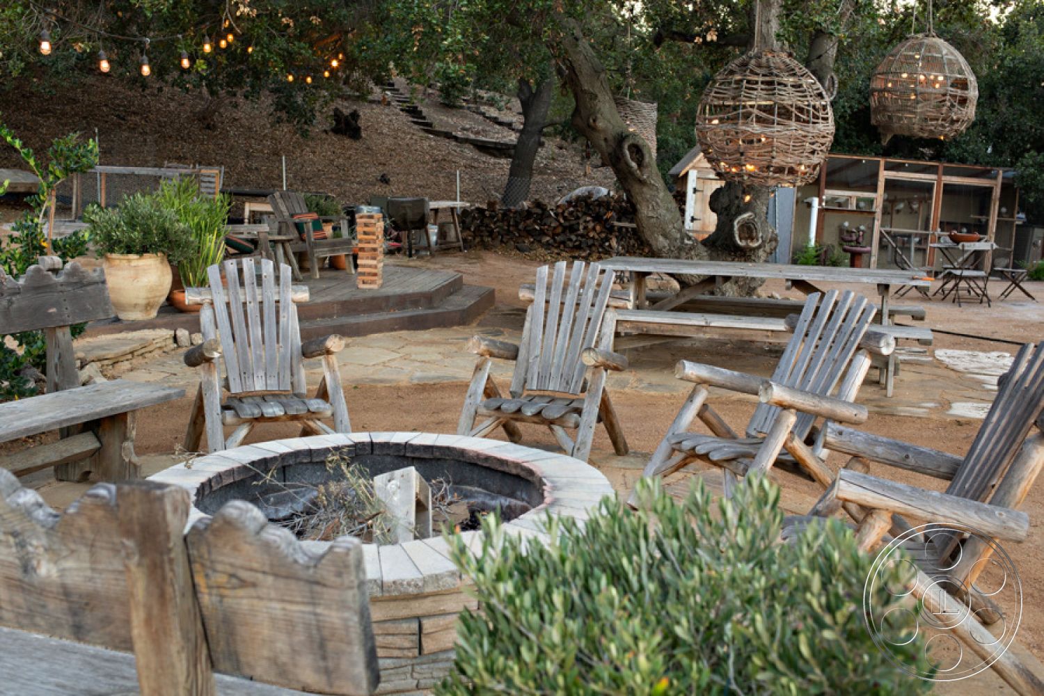 Ranch 33 - wooden picnic table, rustic wooden chairs, natural stone patio flooring, casual outdoor dining area, landscaped garden with greenery, outdoor ambient lighting, exterior wooden deck seating, rural outdoor fire pit area, hanging wicker light fixtures, cobblestone fire pit surround, open-air rustic gathering space, surrounded by trees and natural foliage, informal seating area under trees, backyard gathering setting, earth-toned outdoor decor, fire pit with seating rings