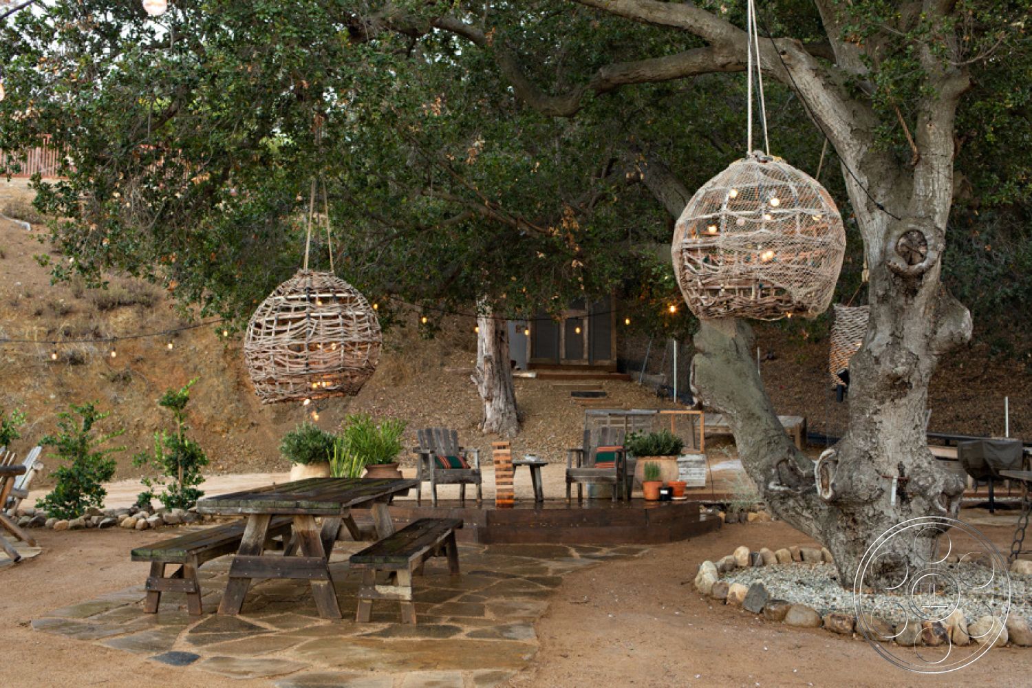 Ranch 33 - exterior, earthy color palette, gravel pathways, landscape lighting, garden setting, hillside location, surrounding oak trees, rustic outdoor seating, open-air gathering area, wood picnic table, textured gravel base, woven hanging light fixtures, natural stone paving patio, natural wood tree branches, decorative rock border