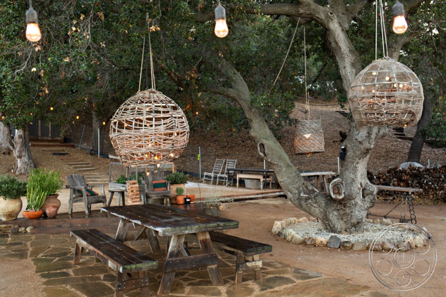 Ranch 33 - hanging light fixtures, outdoor dining area, wooden picnic table, soft ambient lighting, woven pendant lights, reclaimed wood furniture, decorative potted plants, landscaped garden setting, decorative stone border, natural stone patio flooring, rustic outdoor seating, mature oak tree backdrop, earthy soil ground cover, casual outdoor entertaining, backyard gathering space