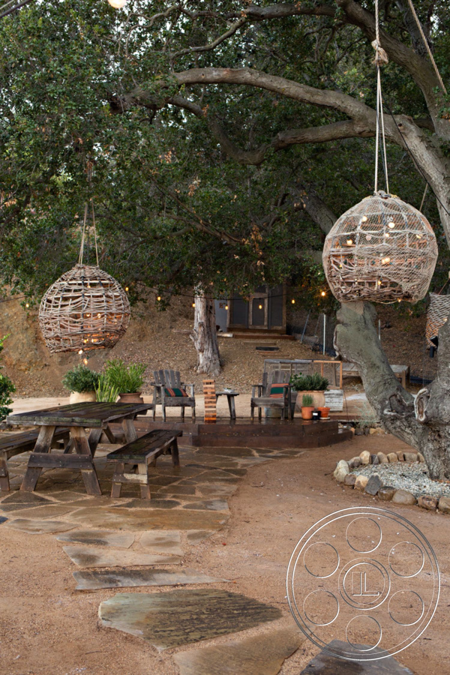 Ranch 33 - ambient outdoor lighting, outdoor lounge chairs, decorative potted plants, tree canopies, landscaped garden beds, exterior patio area, natural stone walkway, rustic garden setting, open-air gathering space, wood picnic table, gravel ground surface, hanging woven light fixtures, wood beam tree support, country-style outdoor seating, wooden furniture finish, peaceful rural environment