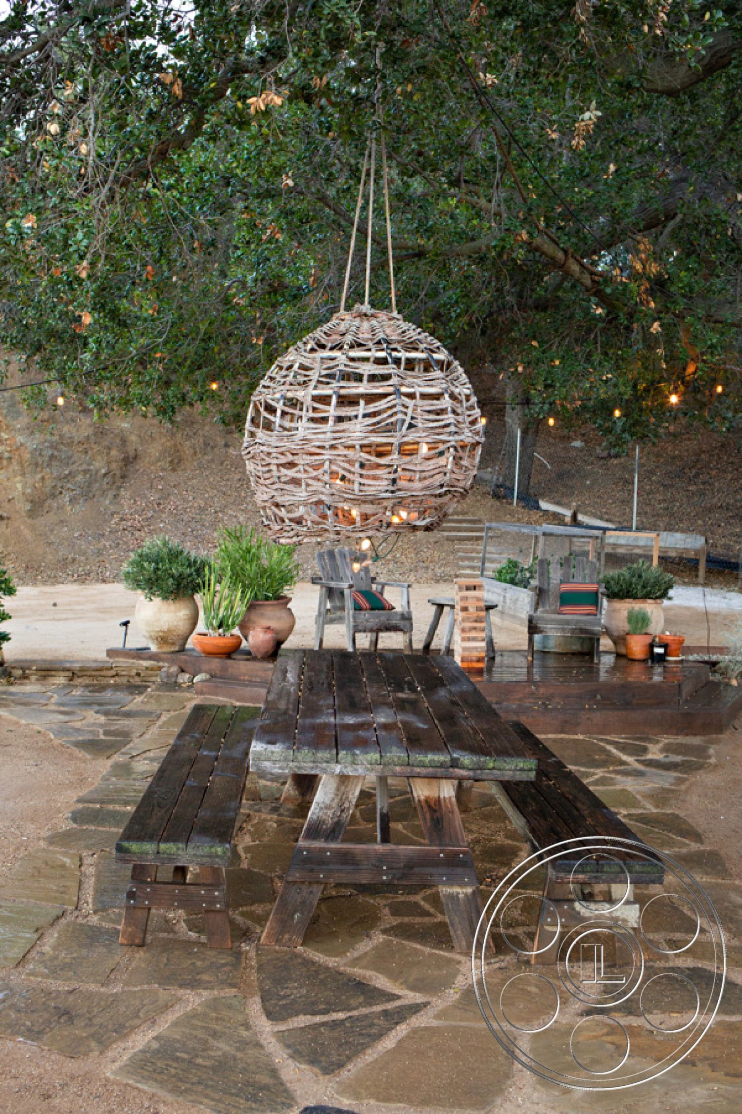 Ranch 33 - casual seating arrangement, woven pendant light fixture, exterior patio area, stone patio flooring, potted greenery accents, surrounding trees for shade, rustic wooden picnic table, weathered wood bench, hanging light decor, natural landscaped setting, relaxed outdoor dining space, textured gravel ground, brick and clay pot planters