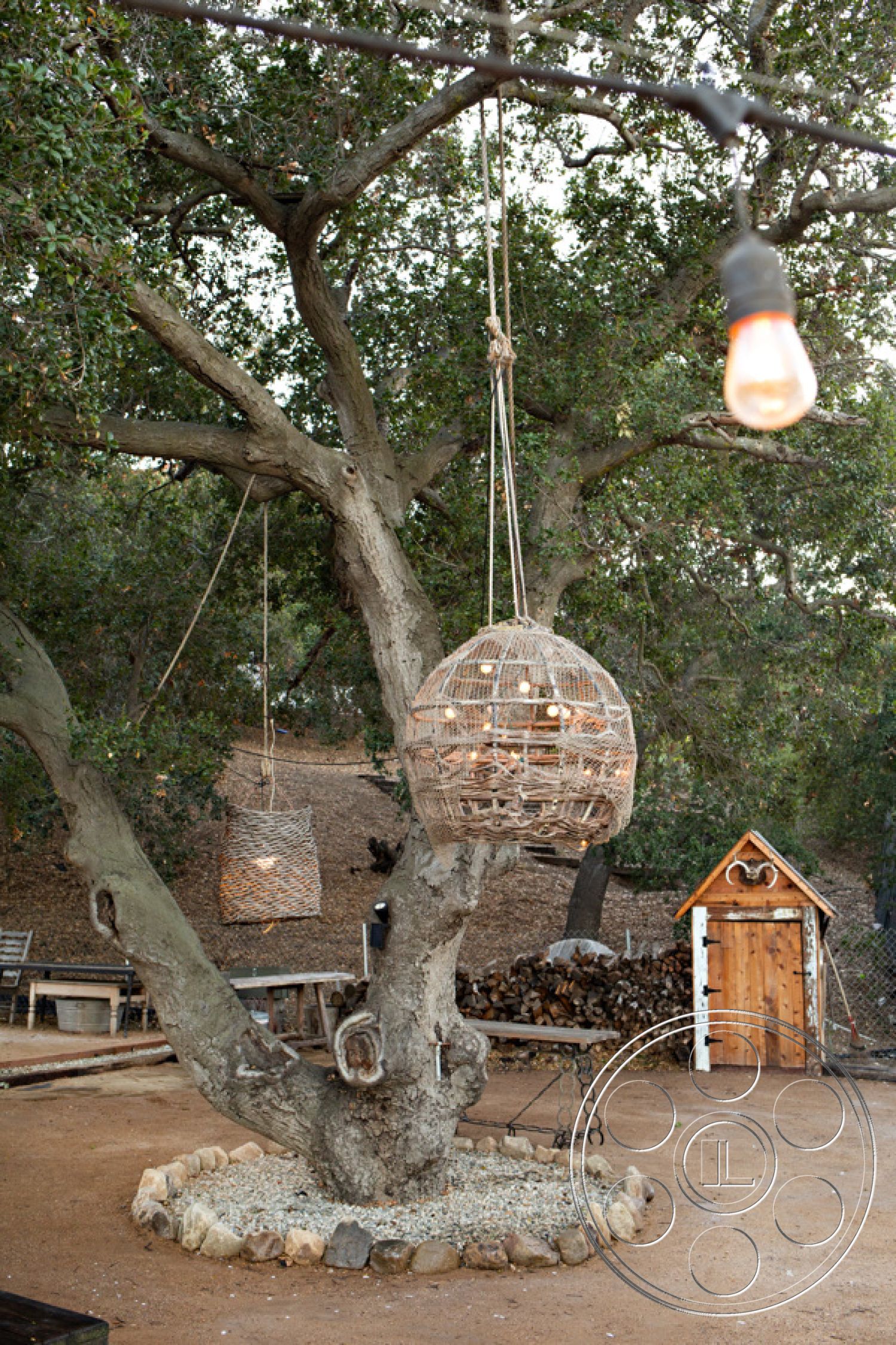 Ranch 33 - outdoor seating area, landscaped backyard, children's play area, open-air setting, cozy gathering space, earth-toned color palette, rustic outdoor decor, exterior lighting fixtures, woven basket pendant lights, rustic tree swing, natural wood tree trunk, gravel ground area, stone border around tree base, wooden shed with a triangular roof, hanging light bulbs, wooden fence in background, tree canopies providing shade