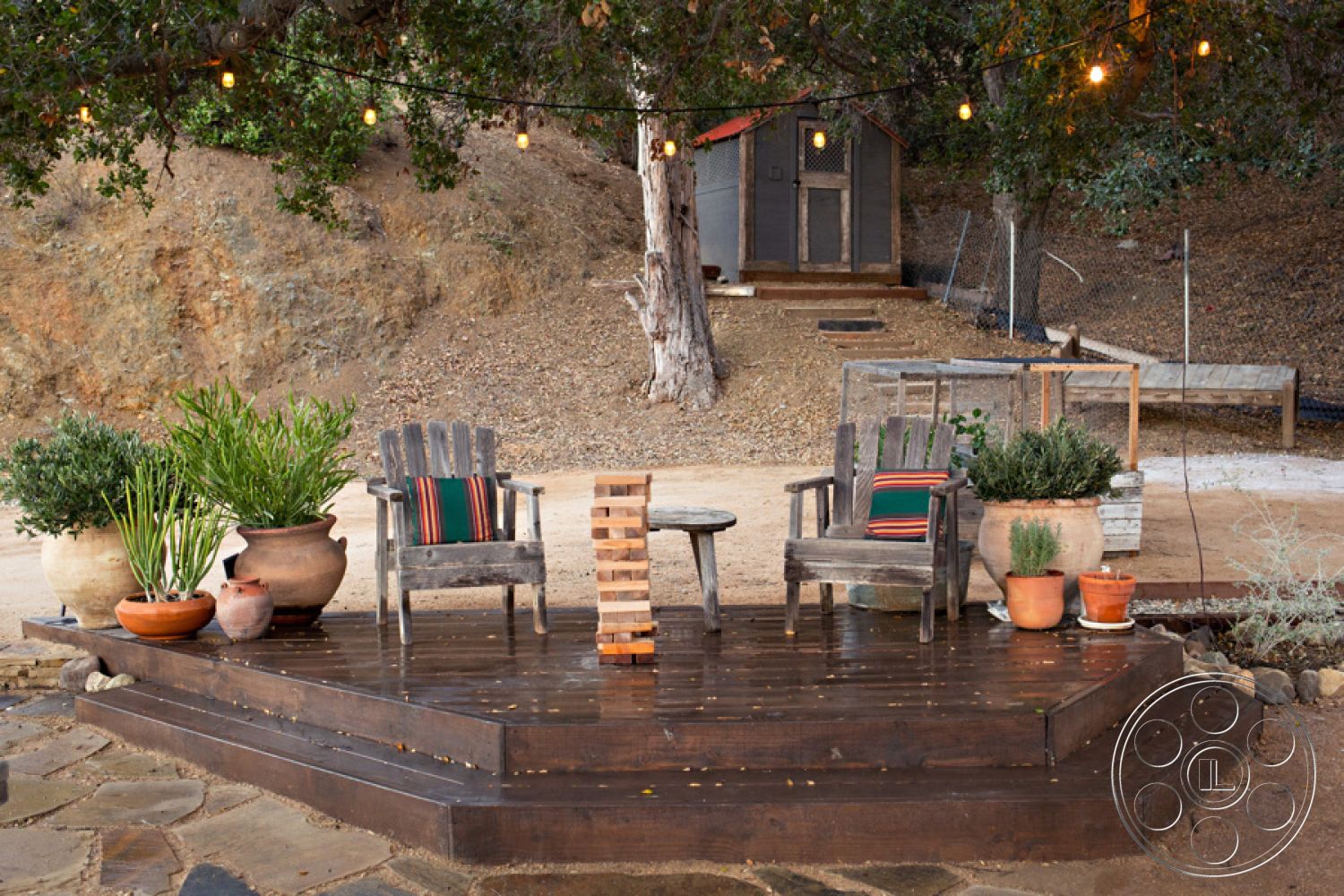 Ranch 33 - stone pathway, natural wood tones, surrounding natural landscape, rustic outdoor seating area, landscaped hillside environment, wooden garden furniture, string light fixtures, weathered wood accents, wooden deck patio floor, rectangular wooden deck, exterior outdoor garden setting, potted plants with terracotta pots, decorative outdoor table, hillside retreat, garden herbs in pots, wooden chair with colorful cushions, cozy outdoor gathering space, exterior rustic decor