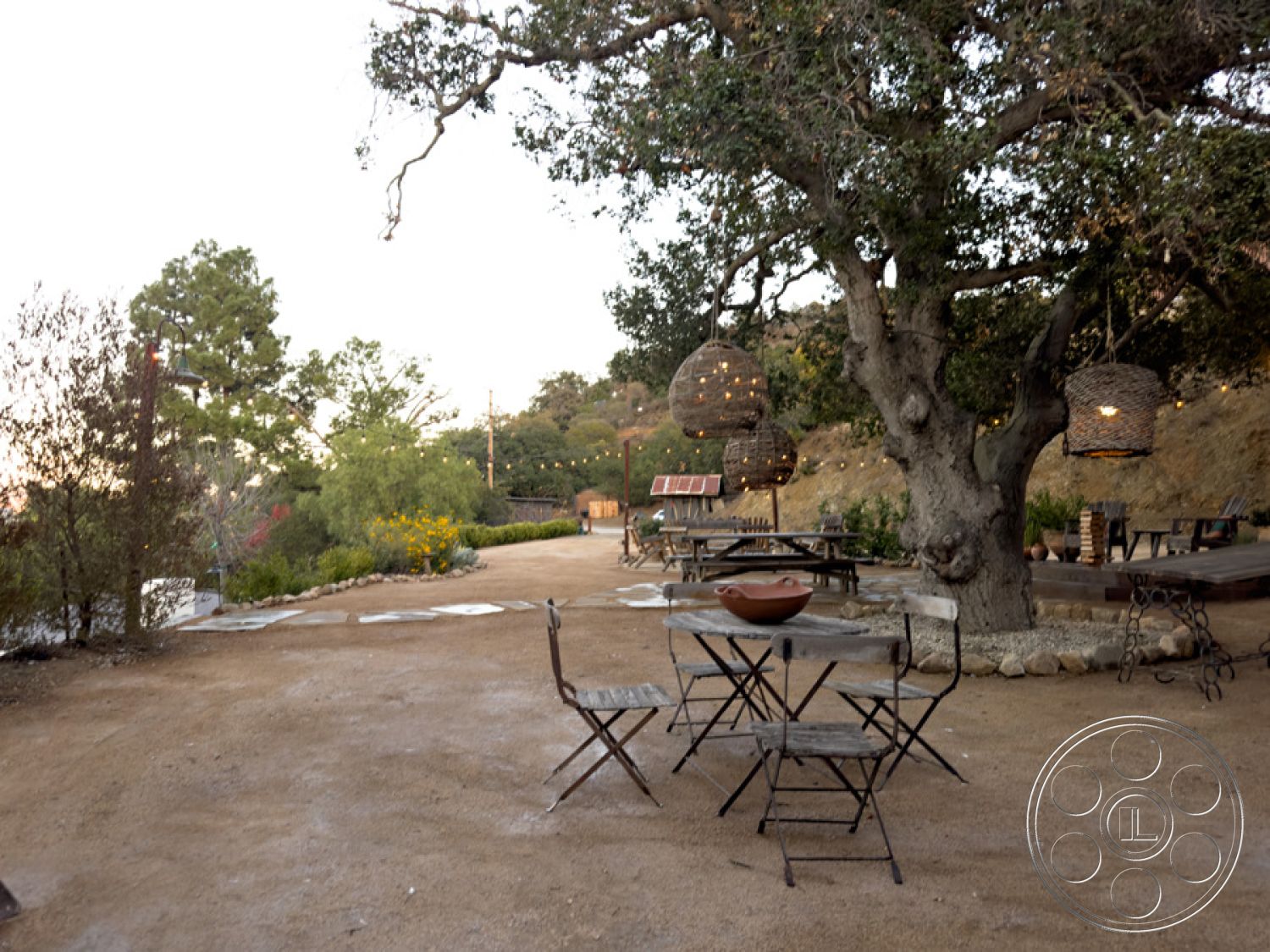 Ranch 33 - exterior, rustic outdoor dining area, natural stone pathway, wooden picnic tables, metal folding chairs, mature oak tree, gravel outdoor flooring, desert landscape setting, landscape with wildflowers, natural elements integration, hanging wicker lanterns, decorative clay bowl centerpiece, light string decorations, patio lounge area, open space with hillside view, casual outdoor entertainment space