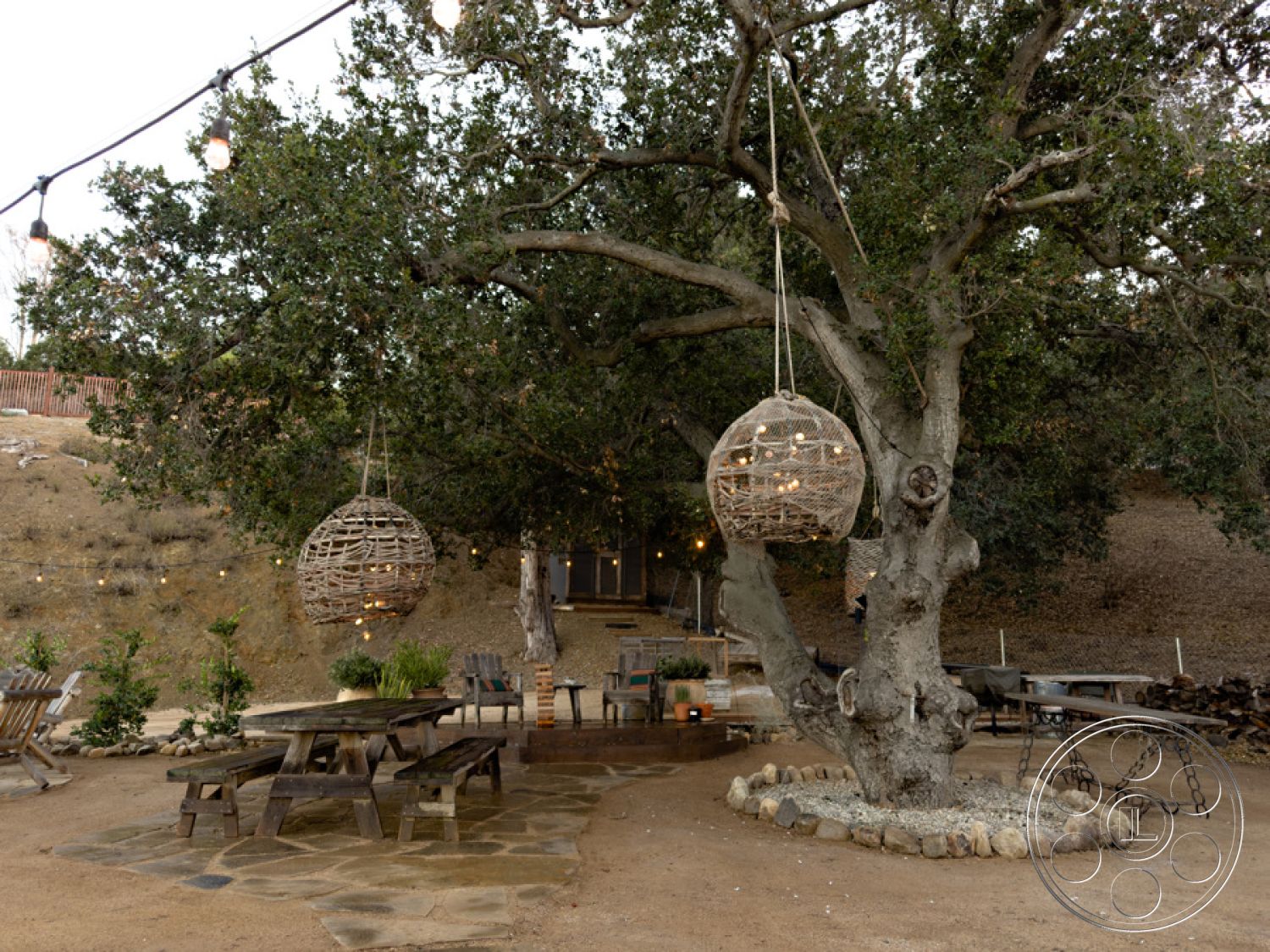 Ranch 33 - exterior, low-maintenance landscaping, outdoor entertaining space, natural stone patio flooring, scenic hillside location, rustic wooden picnic table, hanging wicker lanterns, string lights hung from tree branches, weathered wood tree trunk, country-style outdoor dining area, gravel pathway leading to a rustic home, decorative stone border around trees, mature oak trees providing shade, natural earth tones in outdoor elements, circular stone seating area around the tree