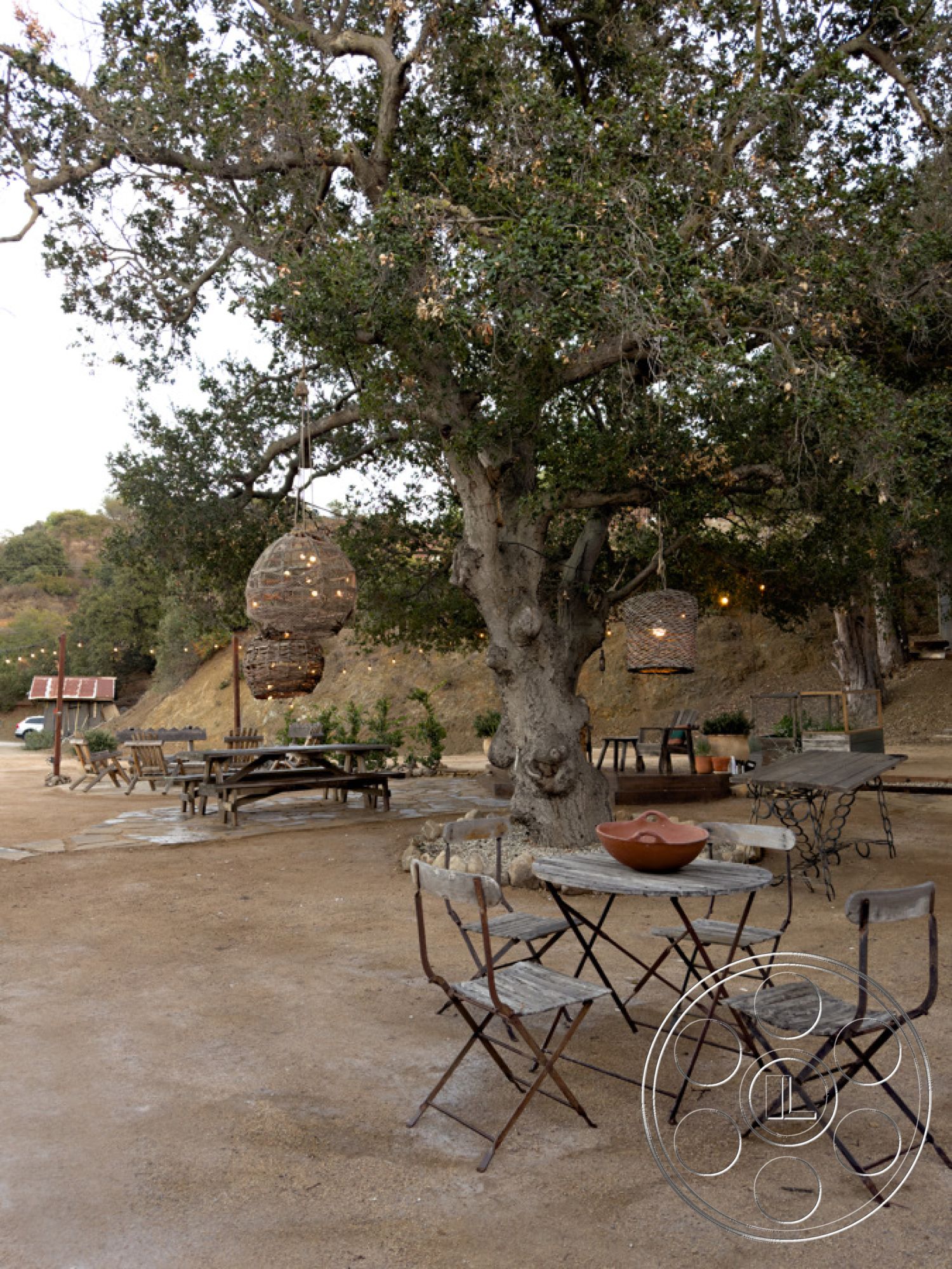 Ranch 33 - exterior, woven pendant lights, hanging string lights, casual gathering space, gravel path, surrounding hills, metal folding chairs, natural stone patio flooring, open outdoor setting, landscaped garden elements, wooden tree canopy, rustic outdoor dining set, earth-toned clay bowl, weathered wooden tables