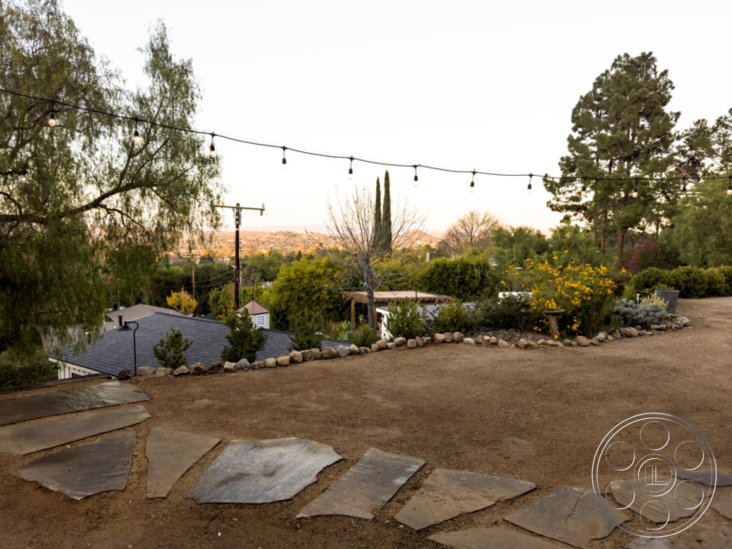 Ranch 33 - exterior, dirt driveway, gravel ground cover, eco-friendly landscaping, native shrub plantings, natural stone patio flooring, mountain view backdrop, irregular stone pathway, open-air outdoor space, landscape lighting with string bulbs, desert-inspired garden setting, gently sloping terrain, circular flower bed with rocks, tree-lined perimeter, attached residence with modern architectural features