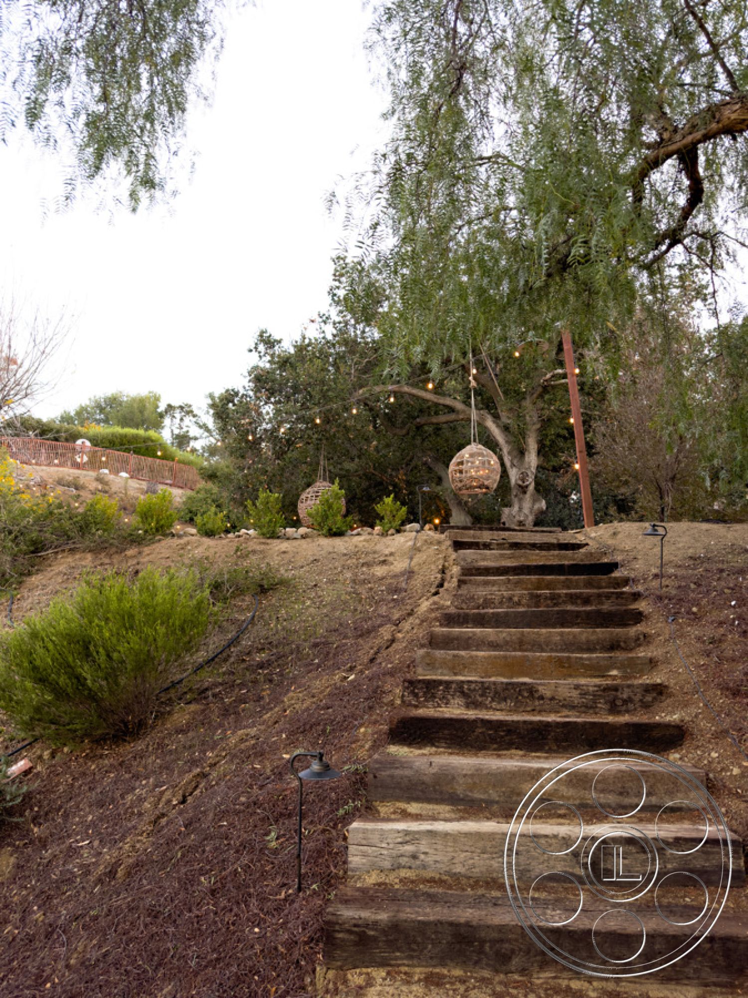 Ranch 33 - landscaped hillside, modern outdoor lighting, mulch ground cover, lush greenery landscaping, natural wood steps, exterior wooden staircase, tranquil outdoor setting, outdoor string lights, decorative hanging lanterns, circular stone accents, rustic natural elements, open garden space, residential hillside pathway, backyard garden retreat, informal outdoor seating area
