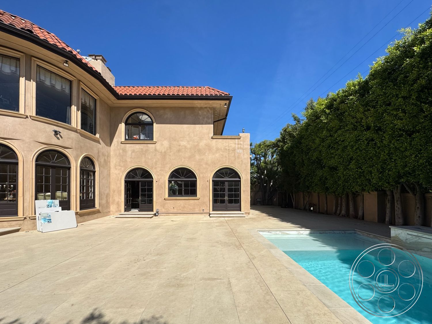 Mansion 146 - exterior, large glass windows, rectangular swimming pool, outdoor living space, landscaped backyard, modern architectural style, wooden fence, arched doorways, concrete patio flooring, stucco exterior walls, open patio area, arced window shapes, clear blue sky view, tile pool border, sunlit backyard, terra cotta tile roof, surrounding hedges