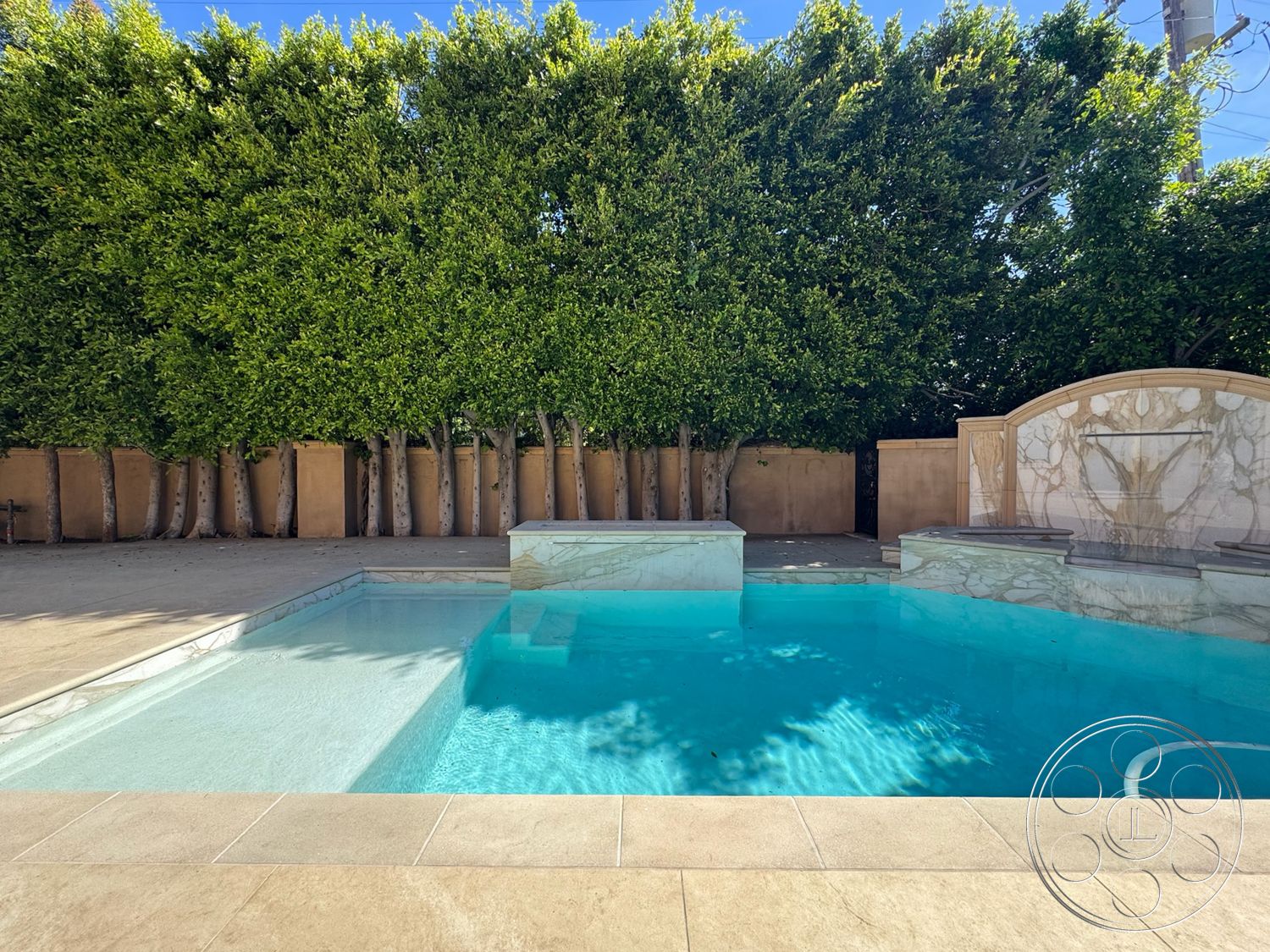 Mansion 146 - modern outdoor space with minimalist design, lush green hedge privacy wall, interior swimming pool with marble water feature, rectangular swimming pool with tiled edge, light beige stone patio flooring around pool, textured plaster wall surrounding pool area, concrete pool steps for easy access, exposed concrete surround with clean lines, sunlit patio space with open sky view, urban backyard pool oasis