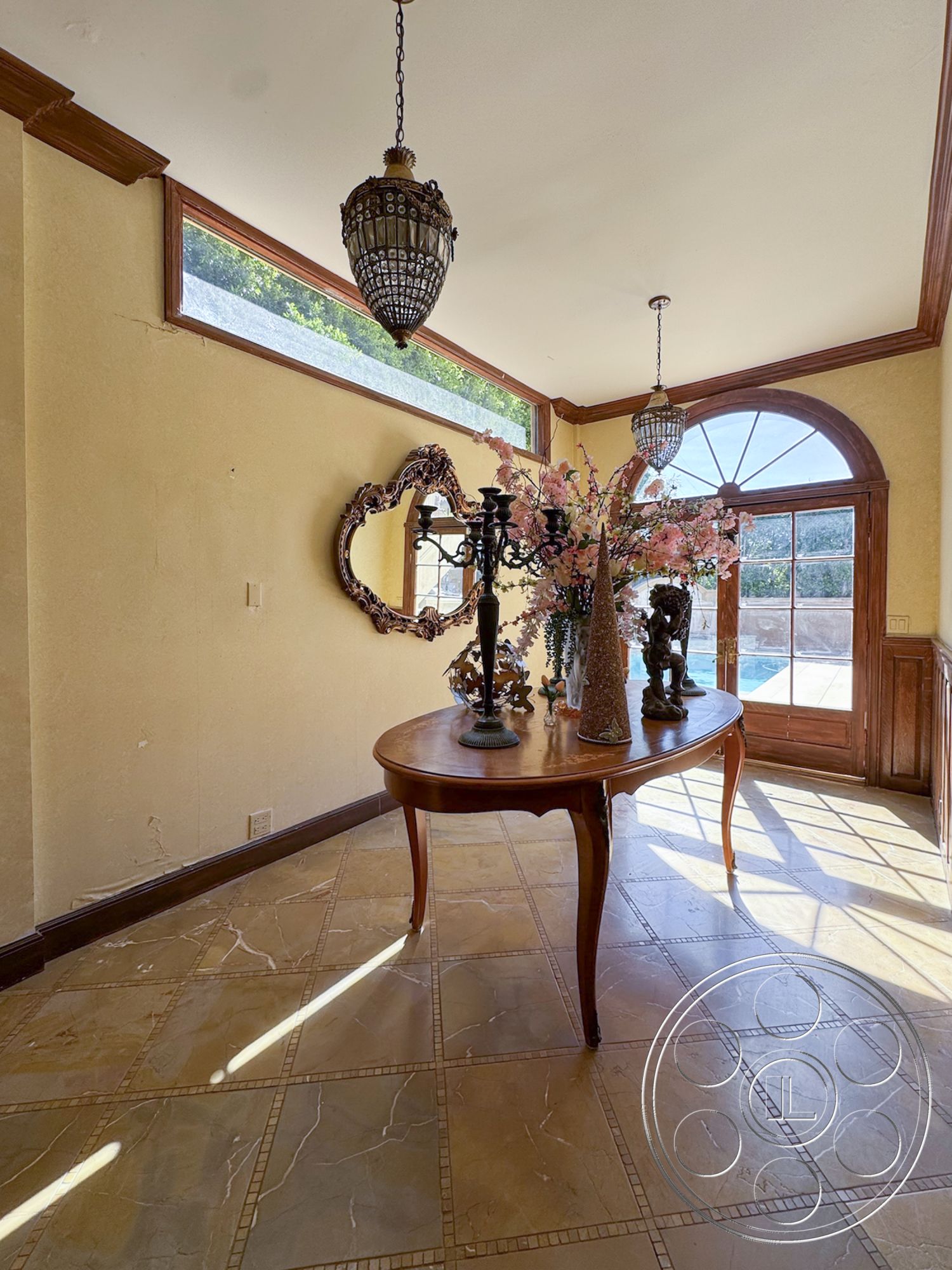 Mansion 146 - natural light from large windows, textured plaster walls, arched entryway, marble entryway flooring, wood trim details, wood panel door, intricate metal chandelier, decorative floral centerpiece, traditional style decor, ornate mirror accent, light-flooded foyer