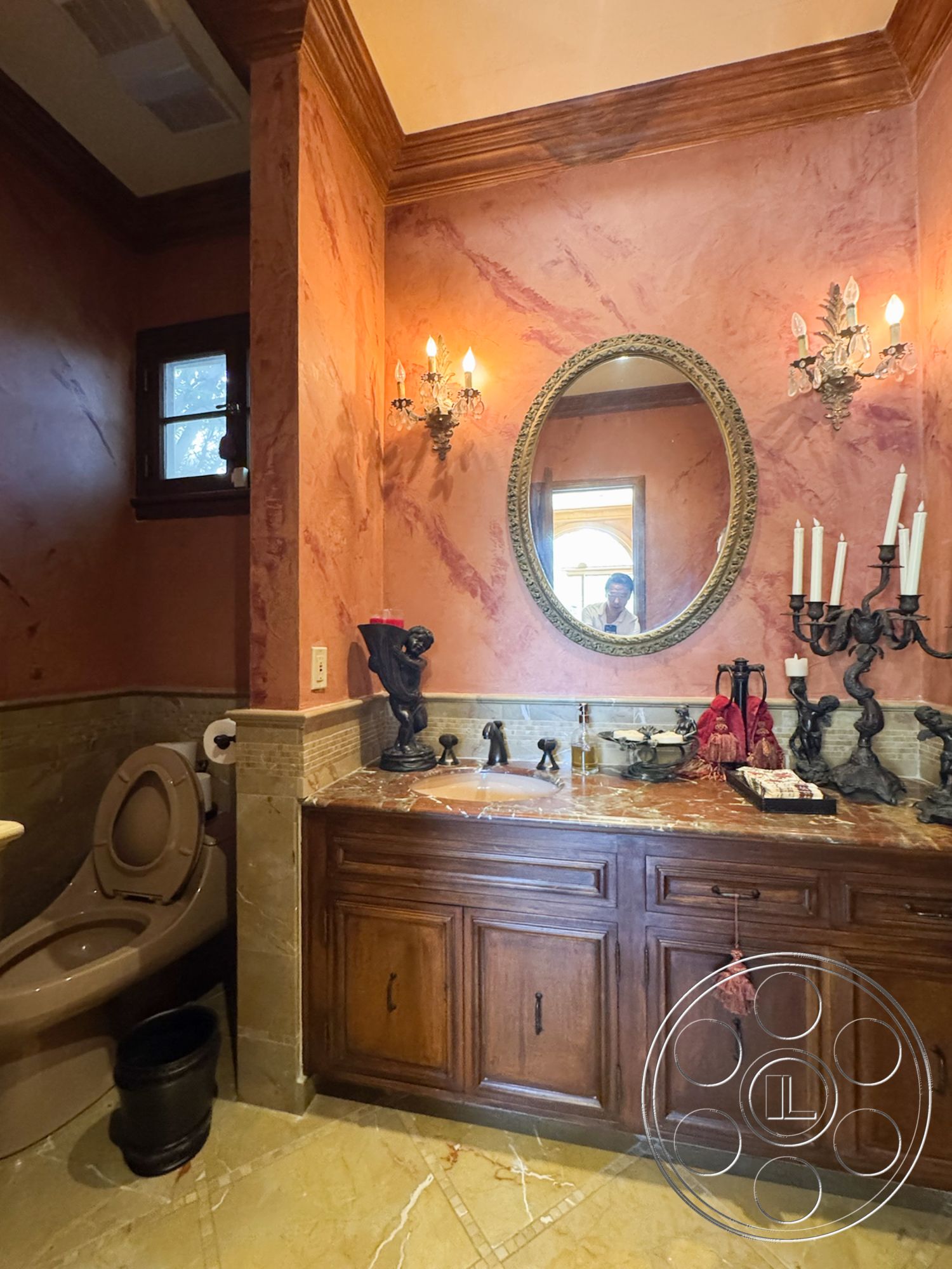 Mansion 146 - high ceilings, decorative candle holders, elegant decor accents, textured plaster walls, traditional wall sconces, cream marble flooring, interior bathroom, small window for natural light, marble countertop sink, dark wood vanity cabinet, pink marbled wall finish, gold rimmed mirror, bronze sink fixtures, wood crown molding, ornate candle stand, classic toilet design