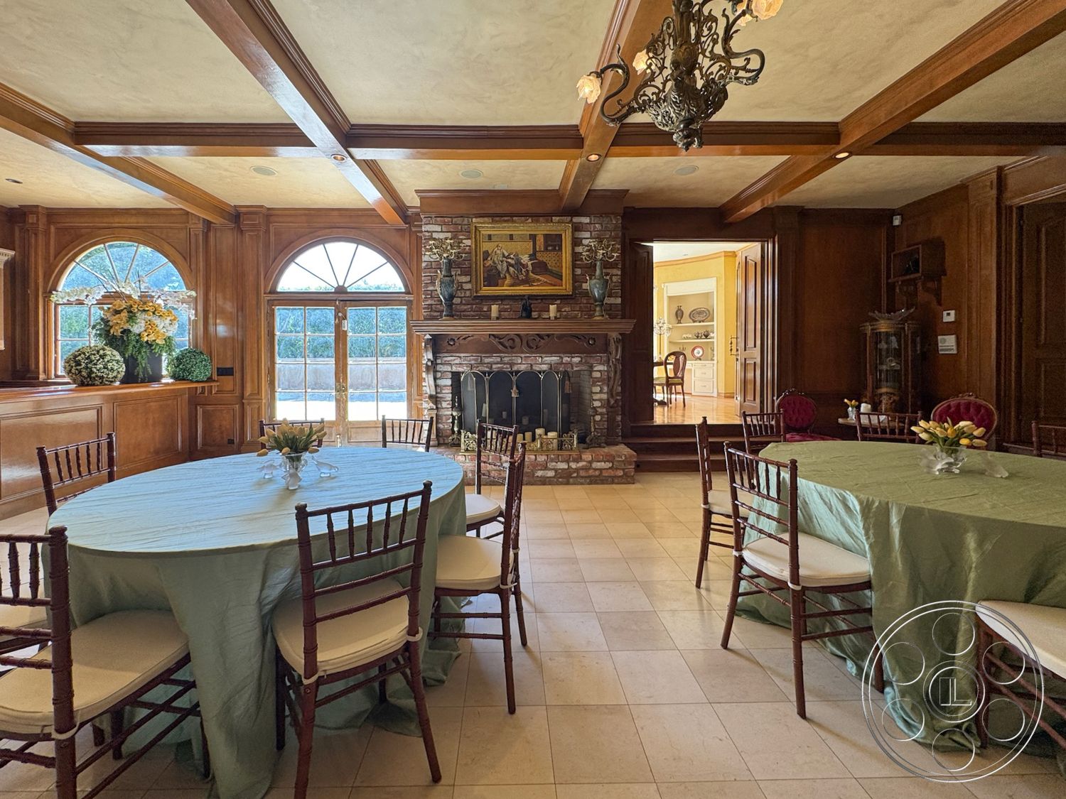 Mansion 146 - neutral color palette, tile flooring, open layout, chandelier lighting, large arched windows, cozy gathering space, stone feature wall, exposed wood ceiling beams, brick fireplace surround, textured plaster wall, oval dining table, hardwood wall paneling, wooden chairs with upholstered seats, elegant table settings